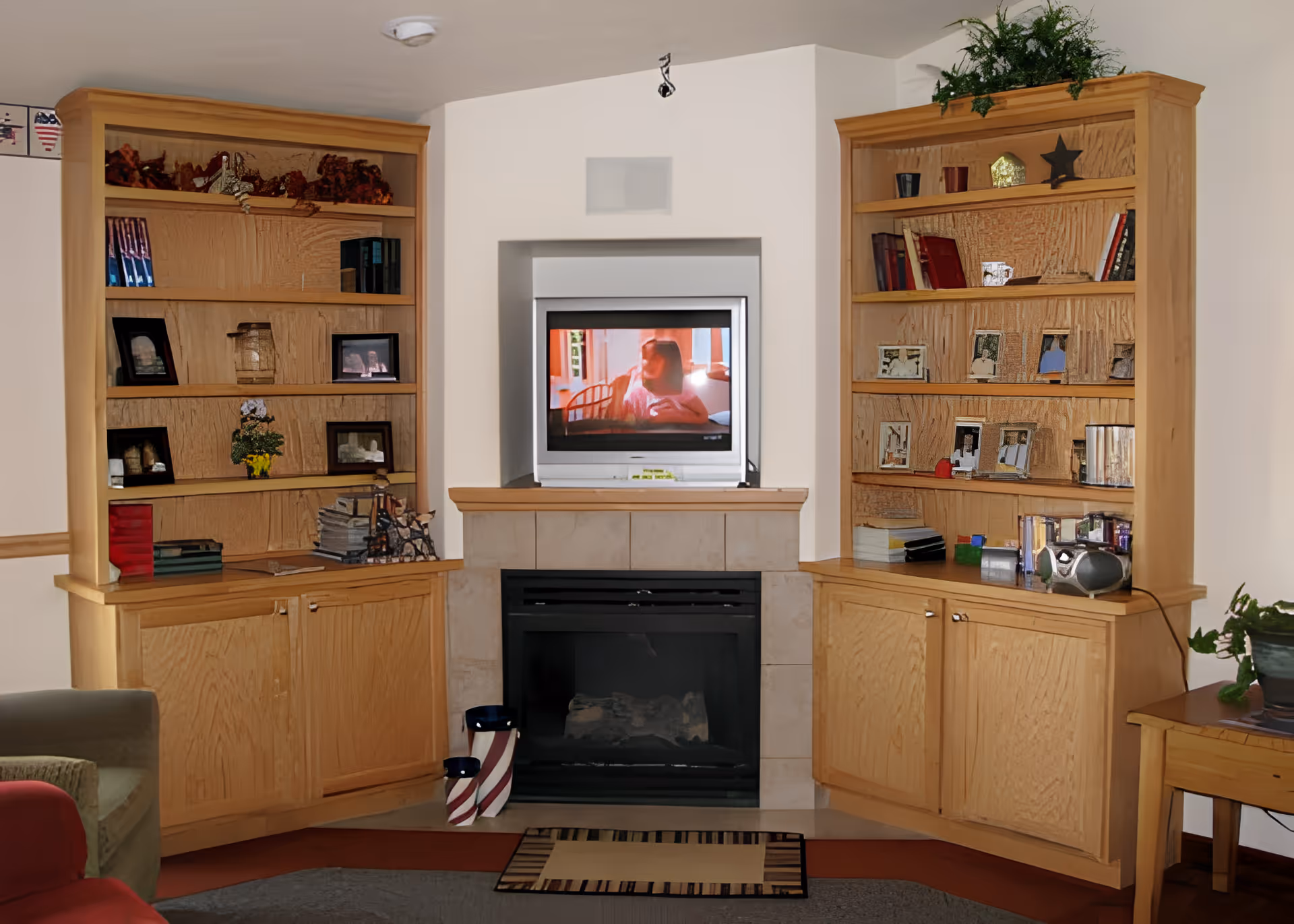 A cozy living room area featuring a fireplace with a TV mounted above it. On either side of the fireplace are wooden built-in shelves and cabinets filled with books, framed photos, and decorative items. There is a small table with a plant on the right side and part of a green armchair visible on the left.