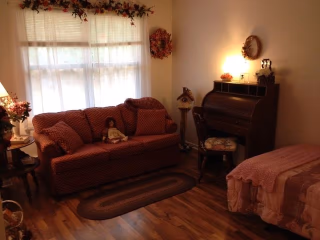 A cozy living space with a red patterned sofa adorned with cushions and a doll, a wooden roll-top desk with a floral cushioned chair, a bed with a pink quilt, a braided rug on wooden flooring, and soft lighting from a lamp on the desk. The window is covered with sheer white curtains and decorated with floral garlands and wreaths.