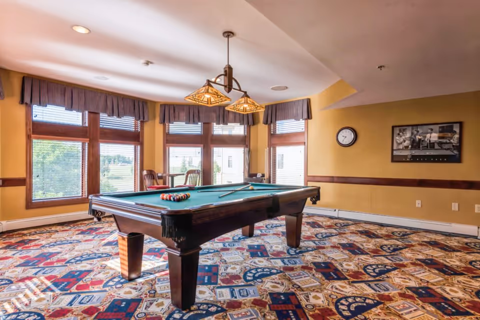 Sunlit recreation room featuring a green-felt pool table on a colorful patterned carpet with large windows and hanging lights.