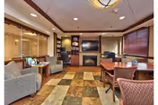 A cozy living room area in a senior living facility featuring a patterned carpet, several upholstered chairs, a wooden table with chairs, a flat-screen TV mounted above a fireplace, built-in shelves with decorative items, and large windows with blinds allowing natural light.