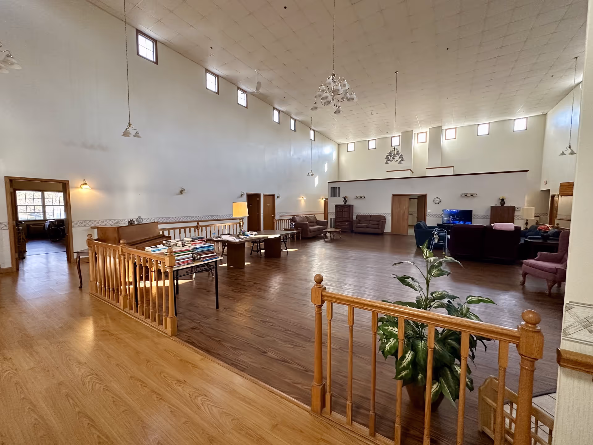 Spacious senior living facility common area with high ceilings and multiple small windows near the ceiling. The room features wooden flooring, several seating areas with sofas and chairs, a table with books, a piano, and a television. There are wooden railings separating different floor levels and a potted plant near the foreground.
