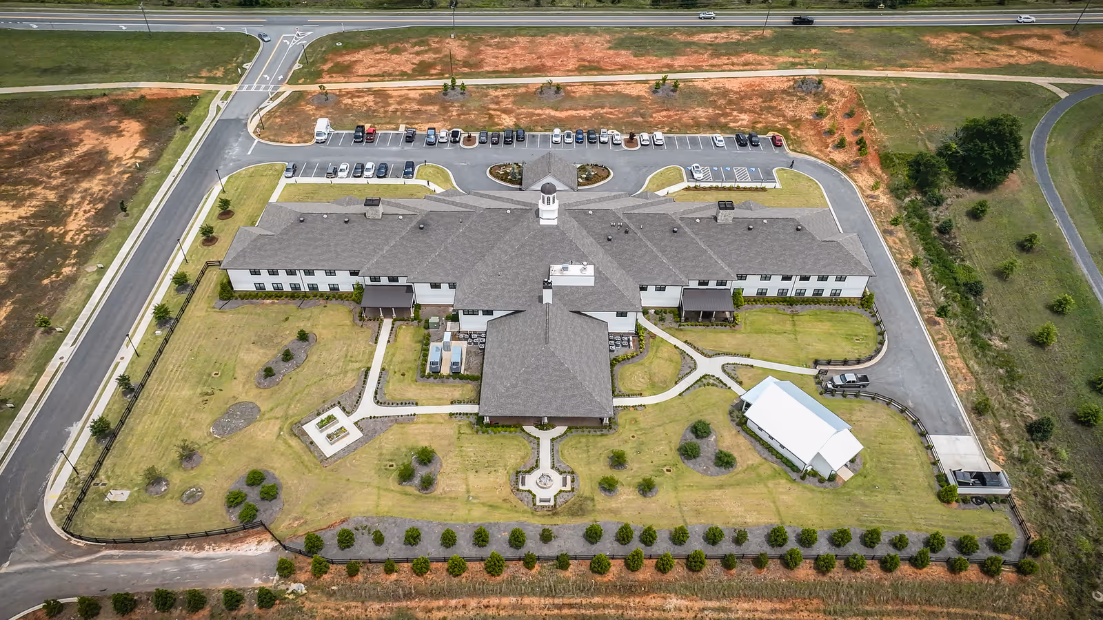 Aerial view of The Fountains in Cartersville assisted living facility showing a large, two-story building with a gray roof surrounded by landscaped lawns, pathways, and parking areas. The building is situated near a road with several cars parked in front. There are small garden areas and a white pavilion on the grounds.