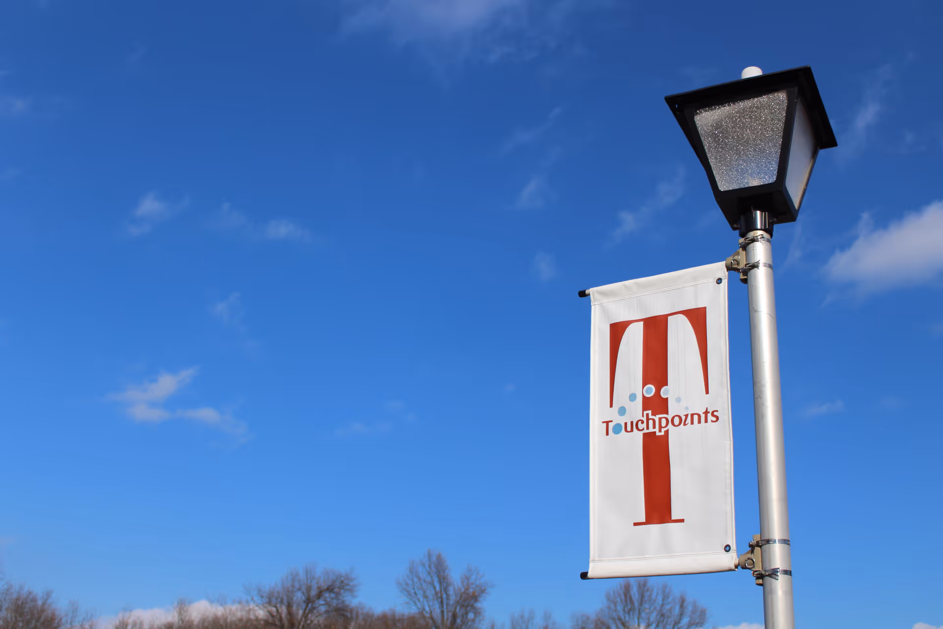 A street lamp post with a white vertical banner attached to it. The banner has a large red letter 'T' and the word 'Touchpoints' written across it. The background shows a clear blue sky with a few clouds and some bare tree branches at the bottom.