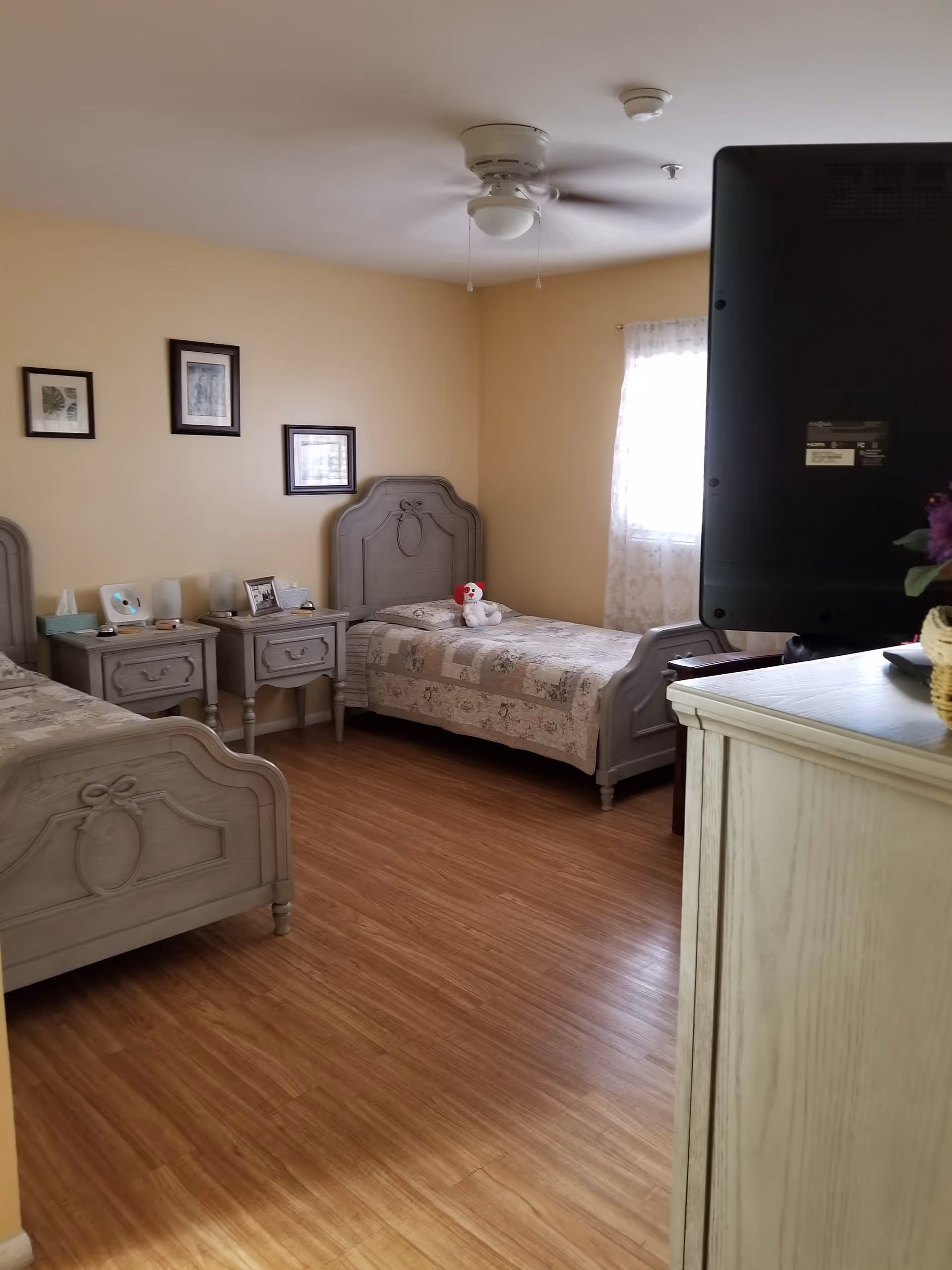 A bedroom with two single beds featuring carved wooden headboards and footboards. Each bed is covered with a patterned quilt. Between the beds are two matching nightstands with lamps, a tissue box, and framed photos. A ceiling fan is mounted on the ceiling, and a window with sheer curtains allows natural light into the room. A TV is partially visible on the right side of the image, placed on a light-colored dresser.