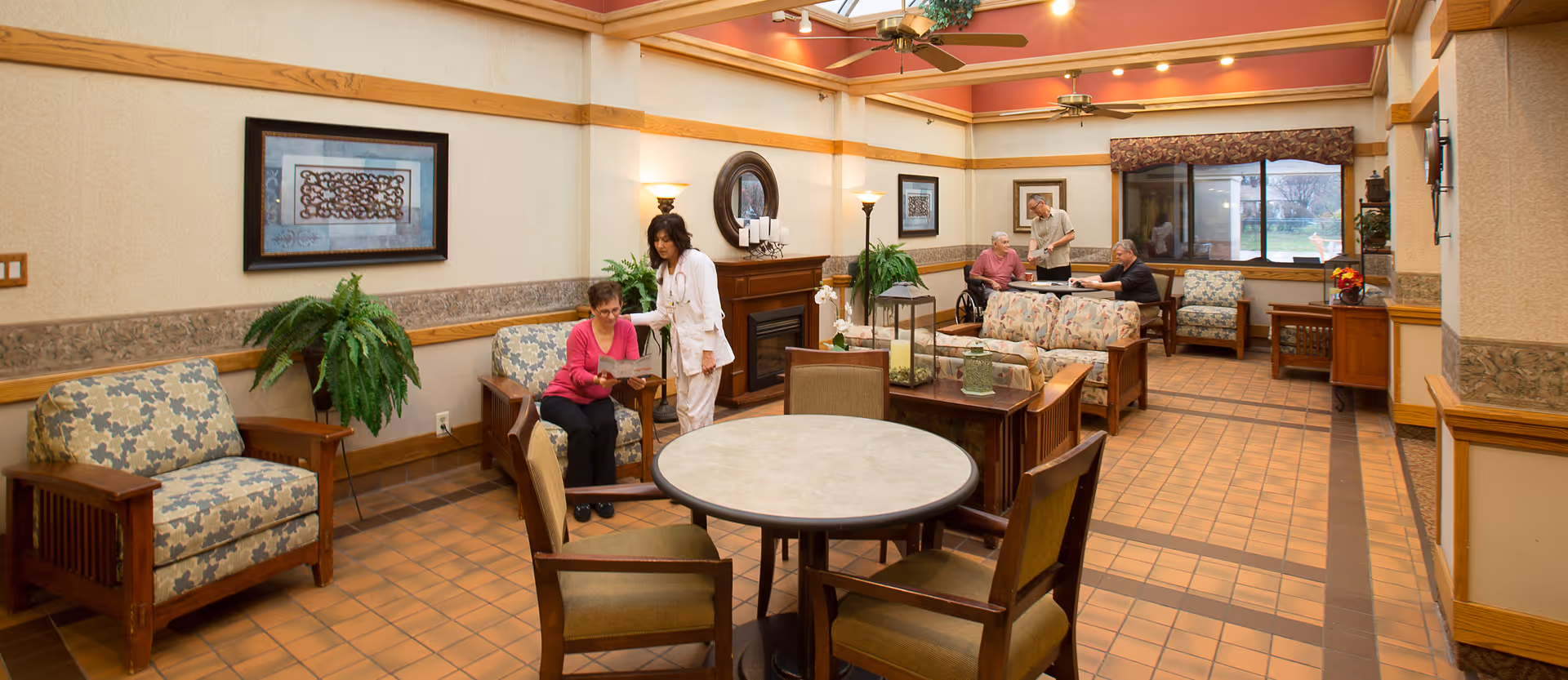 A spacious common area in a senior living facility with several armchairs and sofas arranged around small tables. Two women are interacting near a fireplace on the left side, while three elderly men are seated and conversing near a window on the right side. The room has warm lighting, ceiling fans, framed artwork on the walls, and large windows letting in natural light.