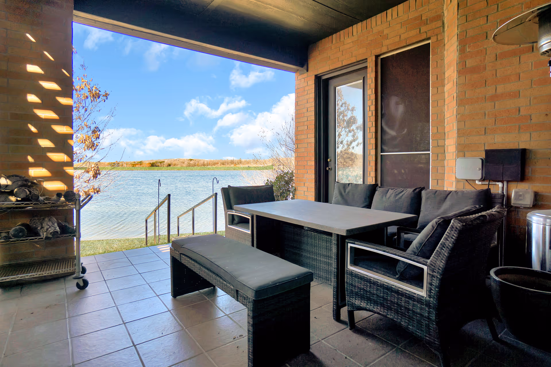 Covered lakeside patio with wicker sofa, chairs, bench and table on a tiled floor overlooking a lake.