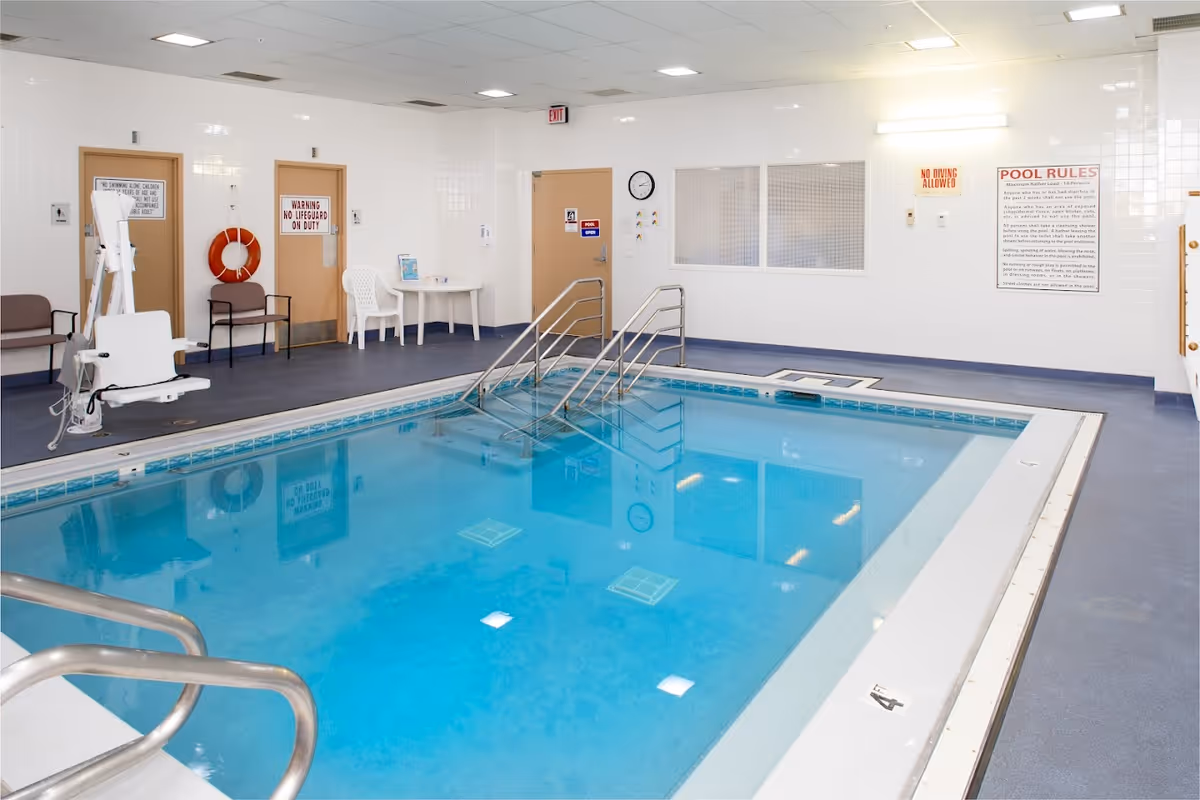 Indoor therapy swimming pool with handrails, a pool lift, seating and pool rules posted on the wall.