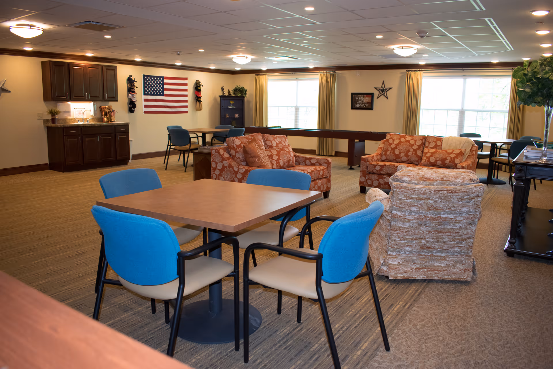 A spacious common area in a senior living facility featuring a table with four blue and beige chairs in the foreground, two patterned orange sofas, and a beige armchair. The room has large windows with yellow curtains, a wall-mounted American flag, a small kitchenette with dark wood cabinets, and a shuffleboard table. The walls are decorated with patriotic-themed items and a star.