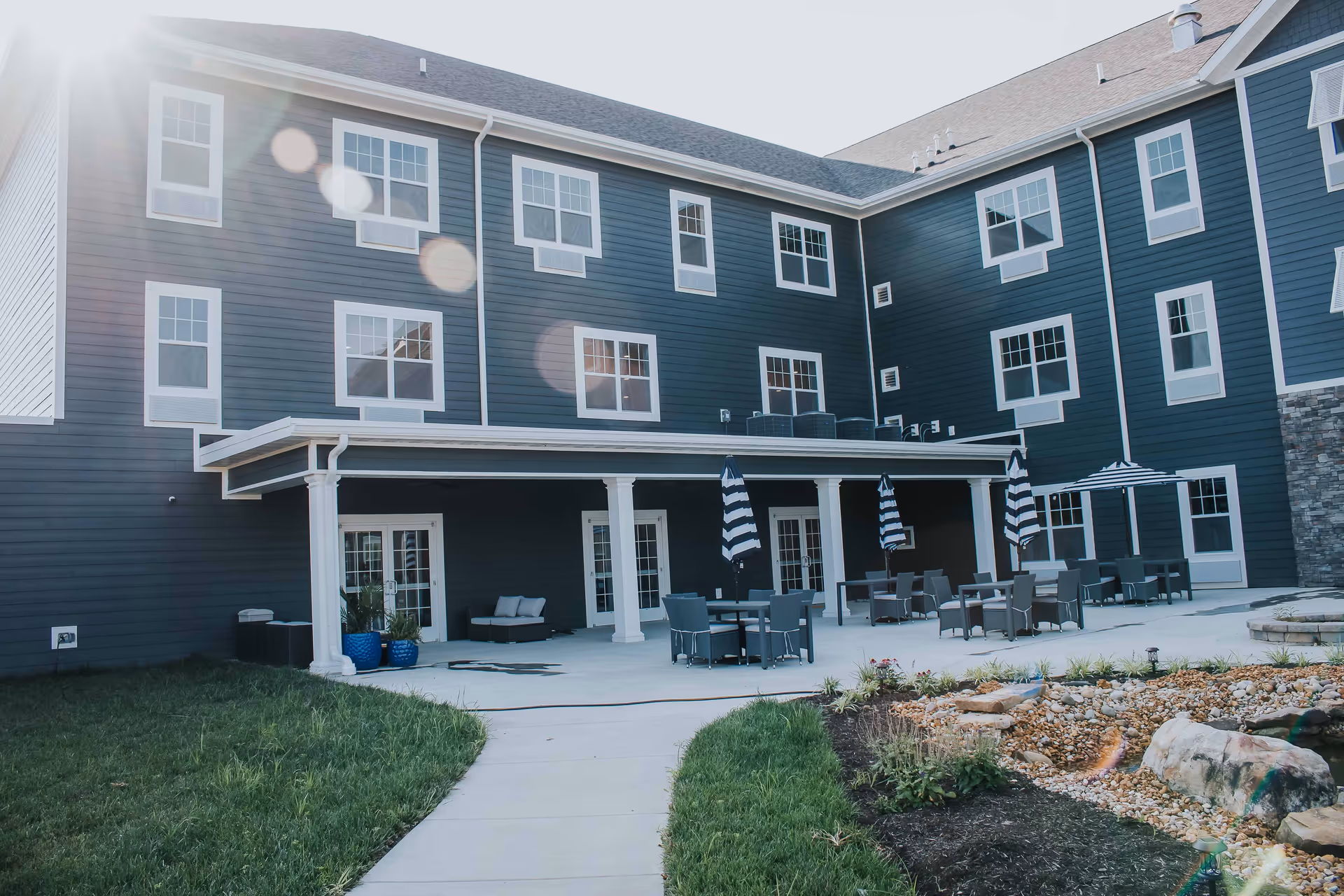 Outdoor patio area of a senior living facility with multiple tables and chairs, some shaded by striped umbrellas. The building exterior is dark blue with white trim and multiple windows. There is a landscaped garden area with rocks and plants in the foreground and a concrete walkway leading to the patio.