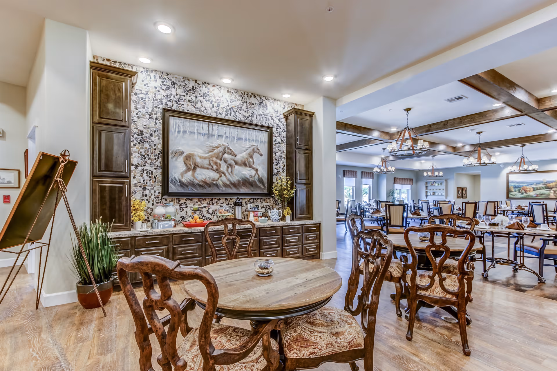 A spacious dining room in an assisted living facility featuring wooden tables and chairs with ornate backs and cushioned seats. The room has wooden flooring, decorative ceiling beams, and multiple chandeliers. A wall with built-in cabinets and a large painting of running horses is visible, along with a small table holding various items and plants.