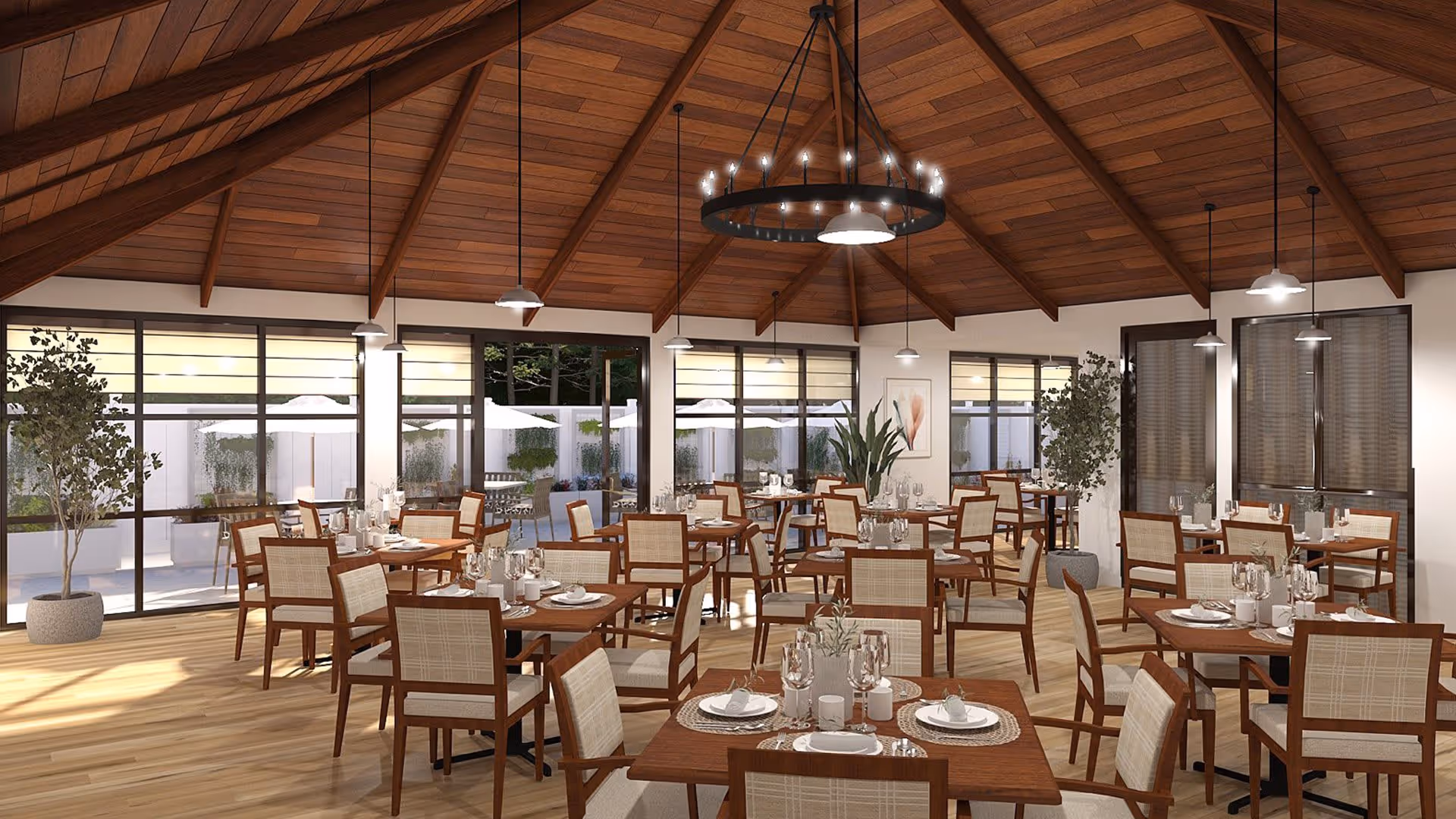 A spacious dining room with multiple wooden tables and chairs arranged neatly. Each table is set with plates, glasses, and cutlery. The room features large windows allowing natural light to enter and providing a view of an outdoor patio area with umbrellas. The ceiling is high with wooden beams and several hanging light fixtures, including a large circular chandelier in the center. There are potted plants placed around the room, adding a touch of greenery.