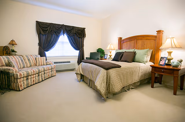 A well-lit bedroom featuring a large wooden bed with beige bedding and multiple pillows, two wooden nightstands with lamps, a framed photo, and decorative items. To the left, there is a striped sofa and a window with dark curtains. The room has beige carpet and light-colored walls.