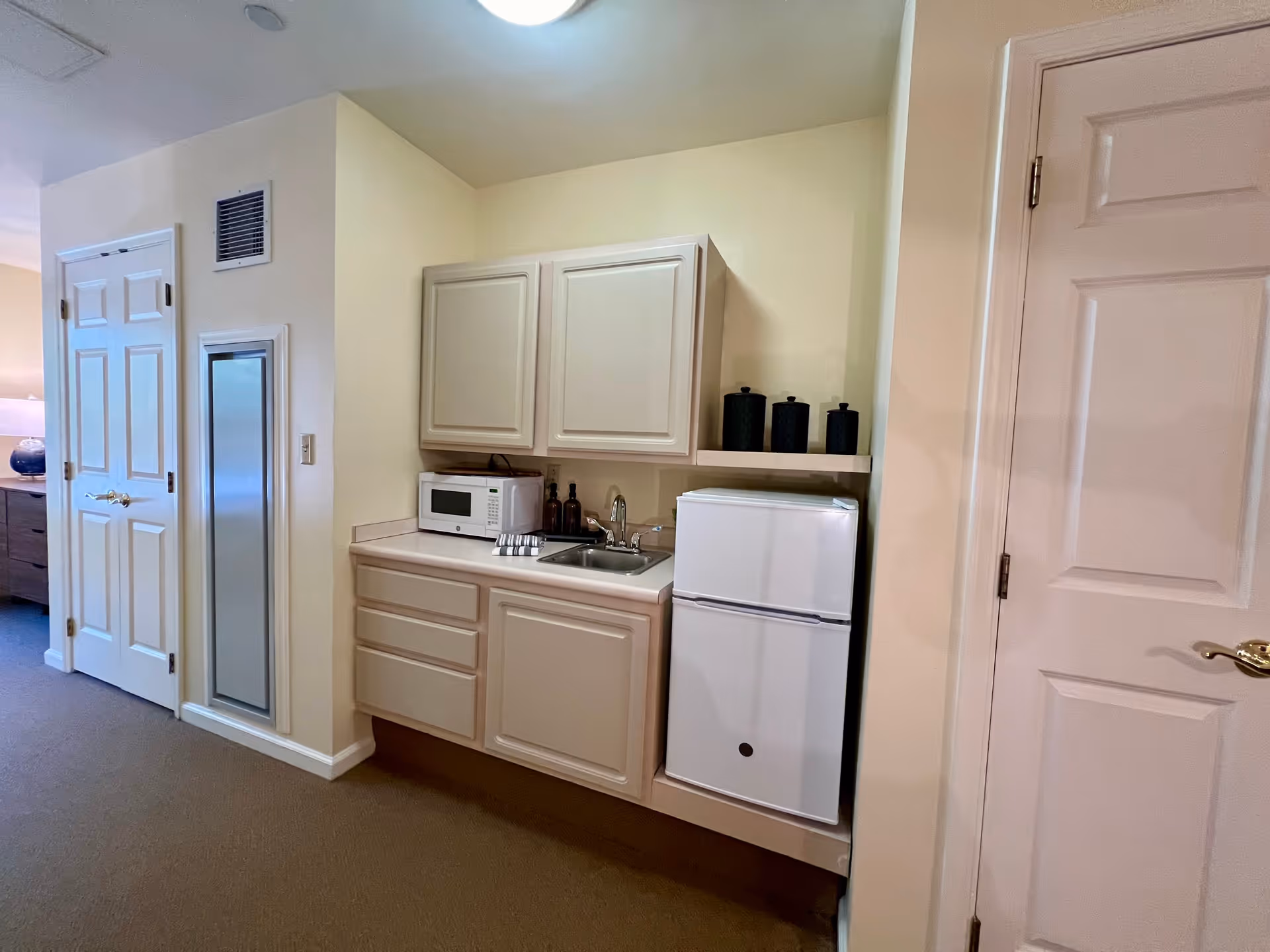 Small kitchenette area with beige cabinets, a white microwave, a small white refrigerator, a sink, and three black canisters on a shelf above the refrigerator. There are two closed doors on either side of the kitchenette, and a carpeted floor.