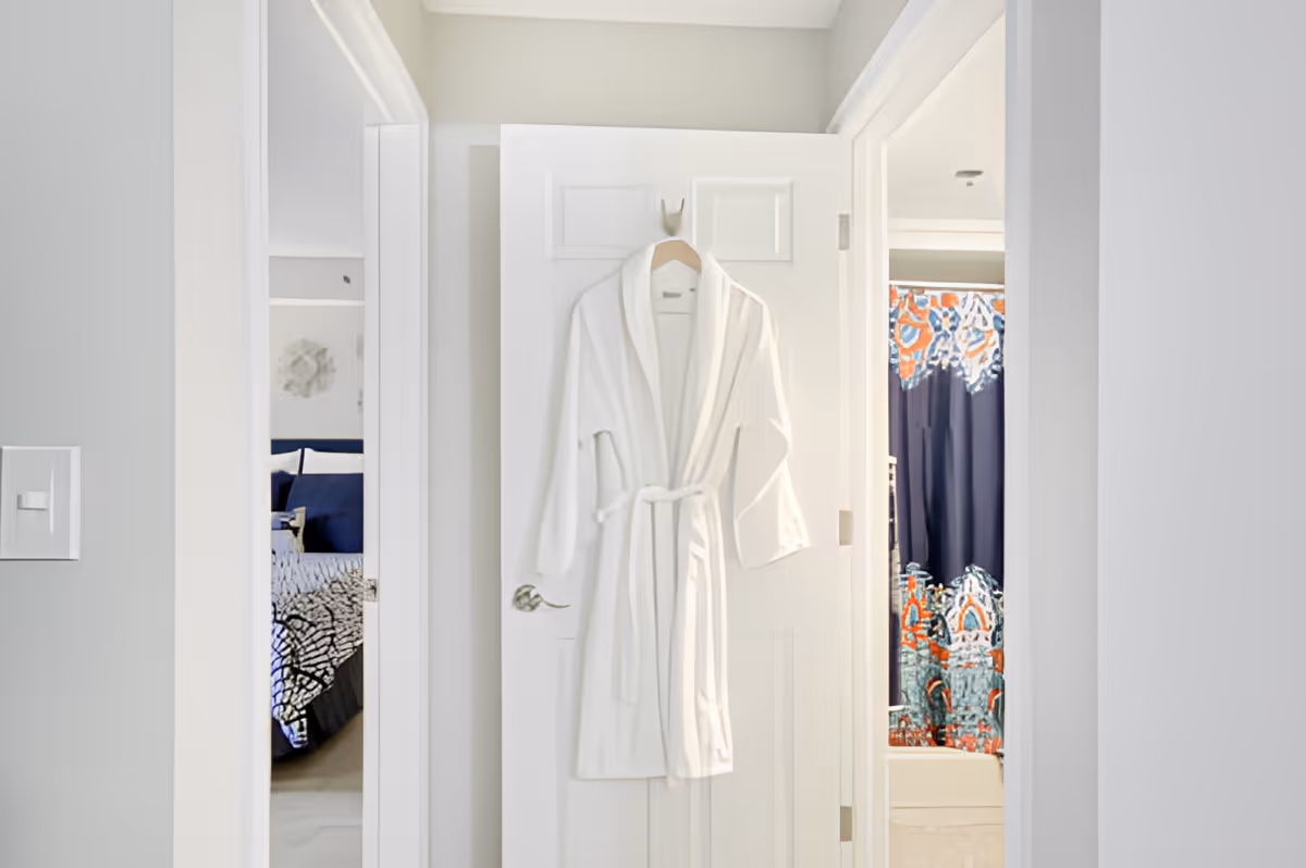 A white bathrobe hanging on a white door in a hallway. To the left, there is a glimpse of a bedroom with a bed covered in patterned bedding and blue pillows. To the right, there is a bathroom with a colorful shower curtain featuring blue, orange, and white patterns.