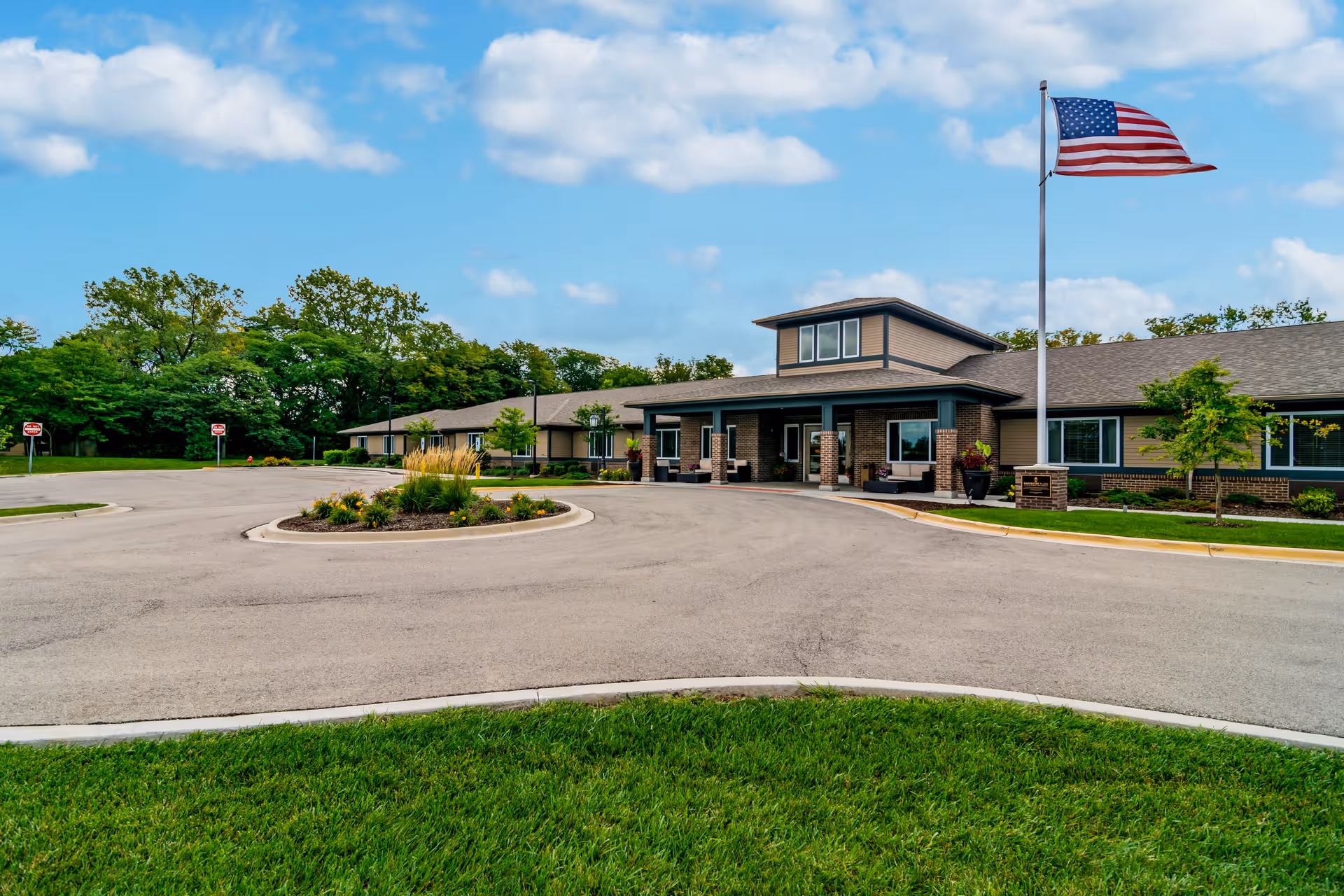 Exterior view of a single-story senior living facility building with a covered entrance, an American flag on a flagpole, a circular driveway with landscaped island, and green trees and grass surrounding the area under a partly cloudy sky.