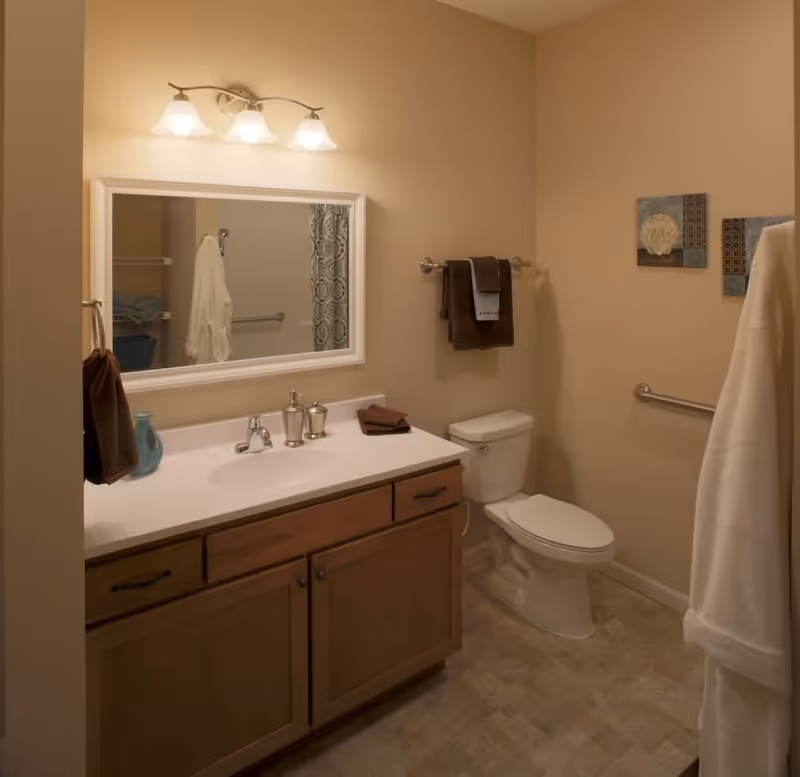 A clean and well-lit bathroom with a white countertop sink, wooden cabinets, a toilet, and beige walls. There is a large mirror above the sink with three light fixtures. Towels hang on the wall and a white robe is visible on the right side. Two decorative wall art pieces are hung on the wall above a grab bar.