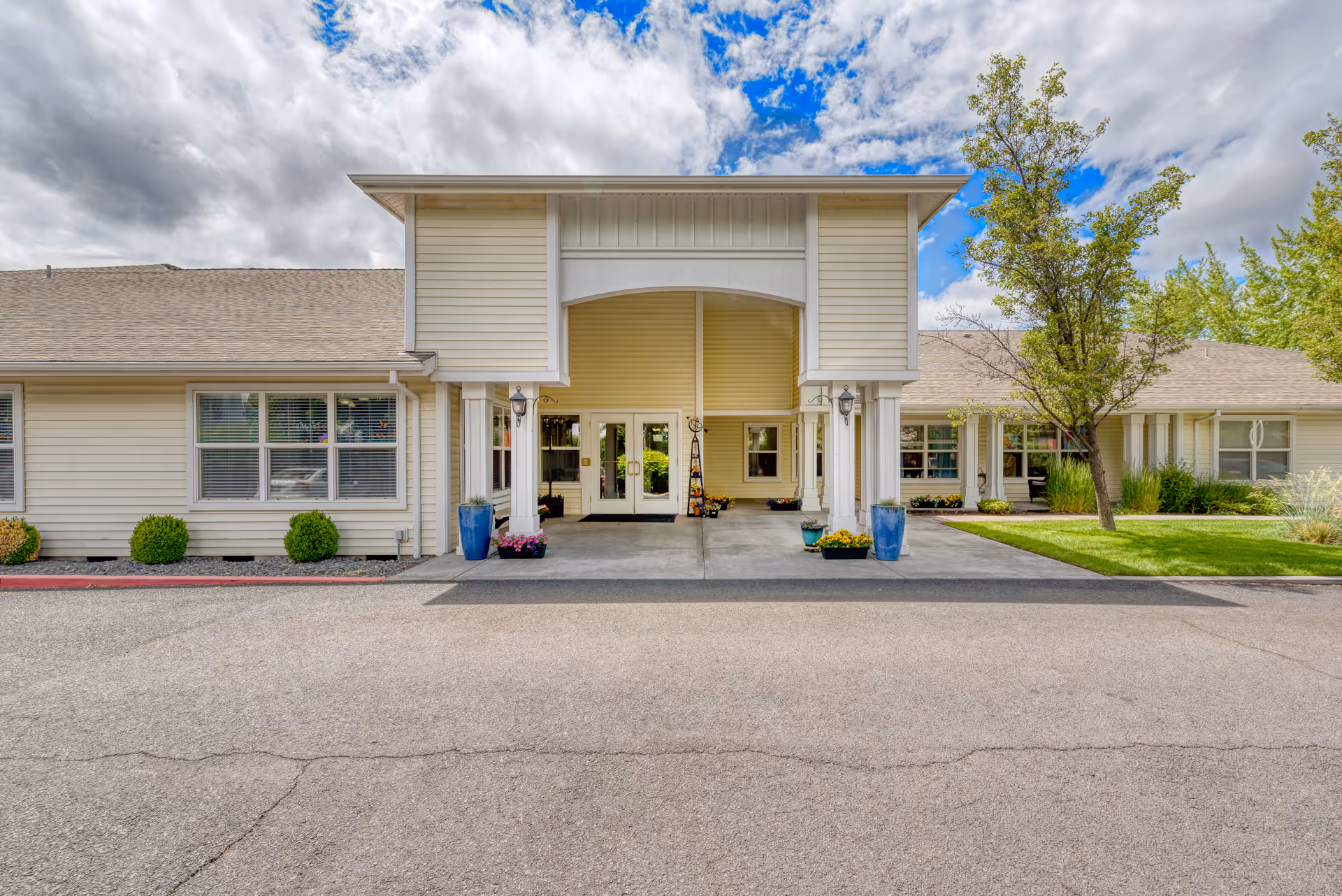 Front exterior view of Awbrey Place Assisted Living and Memory Care building with a covered entrance, beige siding, large windows, potted plants, and a tree on the right side under a partly cloudy sky.