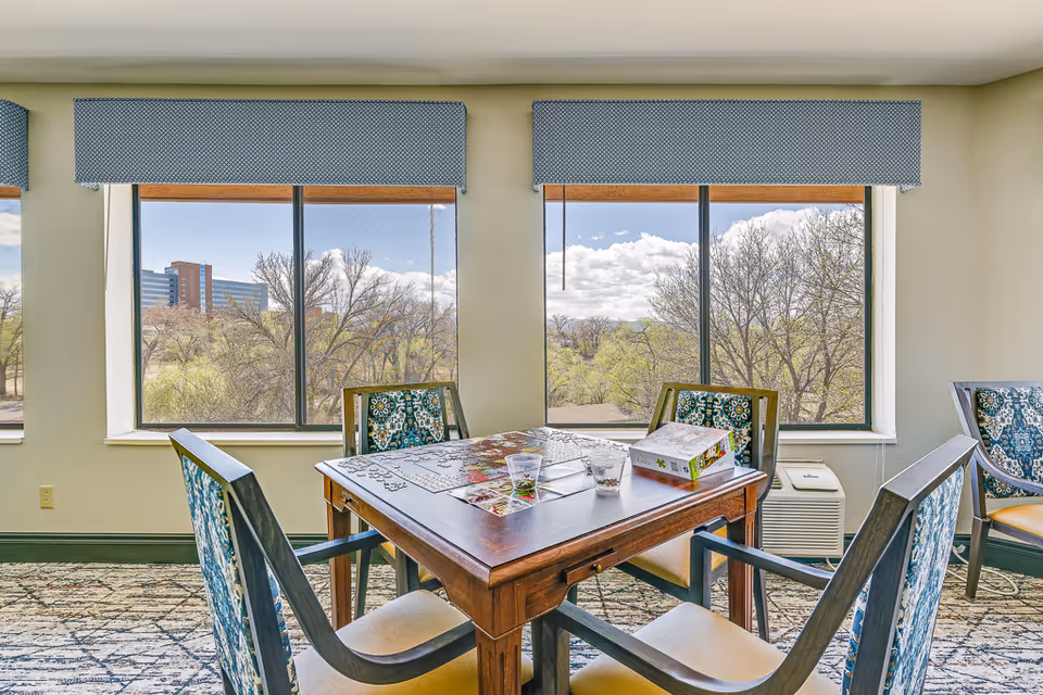 A bright room with large windows showing a view of trees and a distant building. In the center, there is a wooden table with a partially completed jigsaw puzzle and a puzzle box on top. Four chairs with patterned upholstery surround the table. The room has light-colored walls and a patterned carpet.