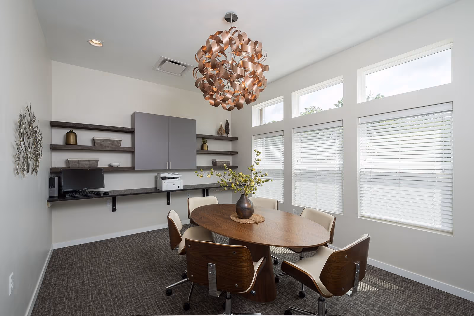 A modern meeting room with a wooden oval table surrounded by six chairs with wooden backs and cream cushions. A decorative vase with green branches is centered on the table. The room has large windows with white blinds letting in natural light. On one wall, there are floating shelves with decorative items, a computer, and a printer. A unique, artistic copper-colored chandelier hangs from the ceiling.