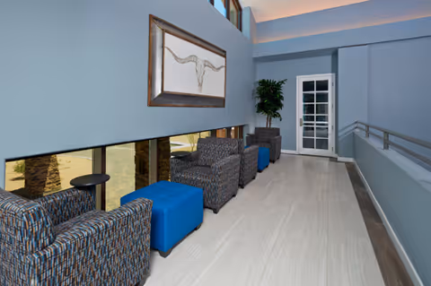 A modern indoor seating area with patterned armchairs and blue ottomans along a light-colored floor. A large framed artwork hangs on the blue wall above the chairs. There is a potted plant near a white door with glass panels, and a railing runs along the right side of the space.