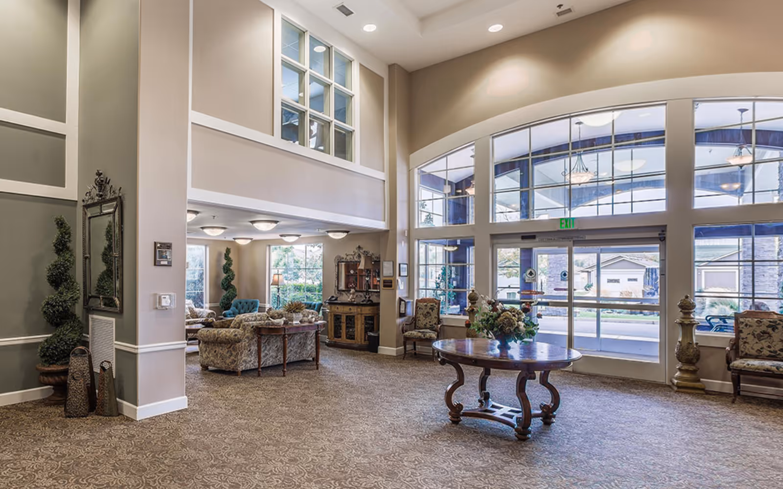 Spacious senior living lobby with large arched windows, seating areas, and a round table topped with a floral arrangement.