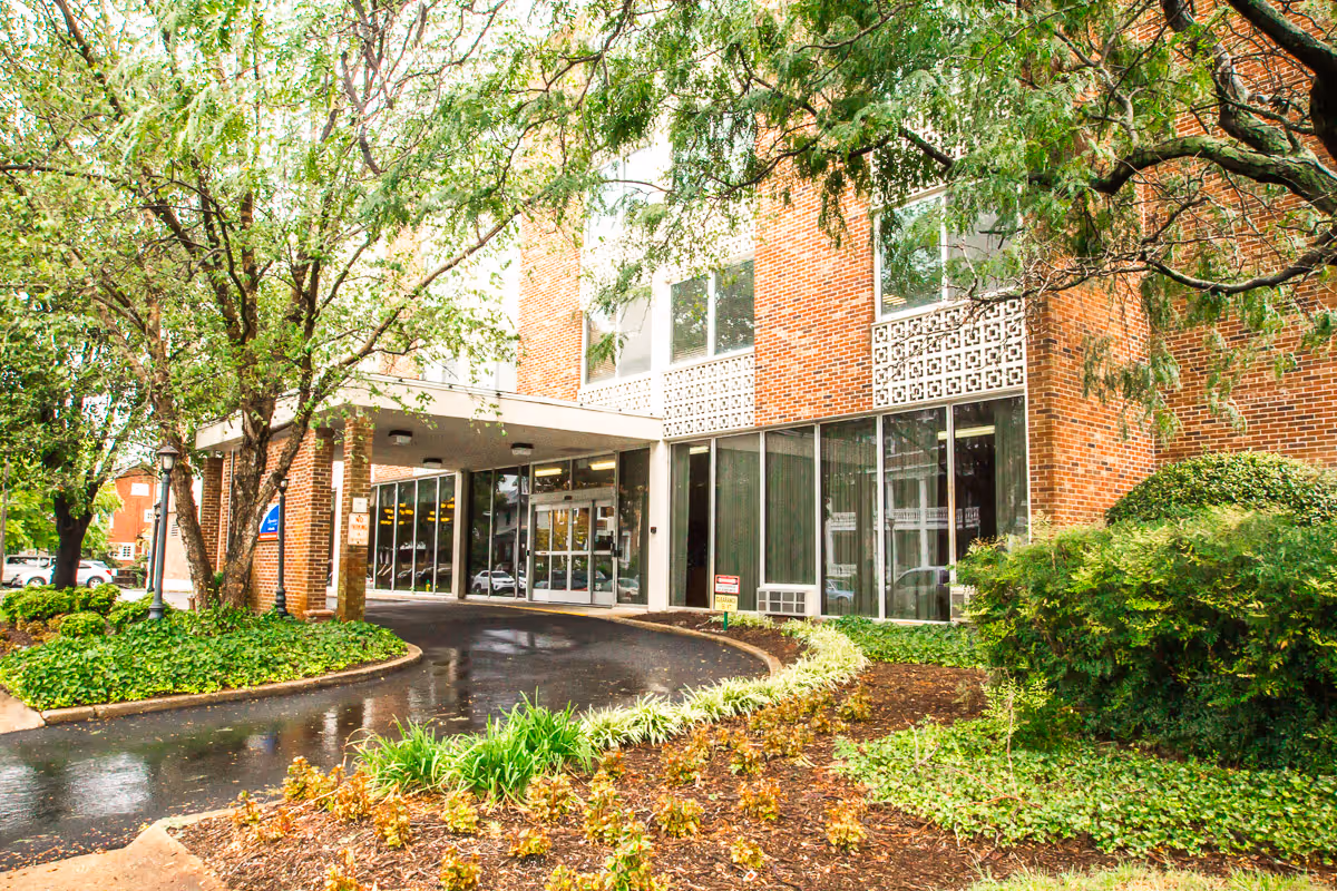 Front entrance of a brick senior living facility with a covered drop-off driveway, glass doors, and surrounding trees and landscaping.