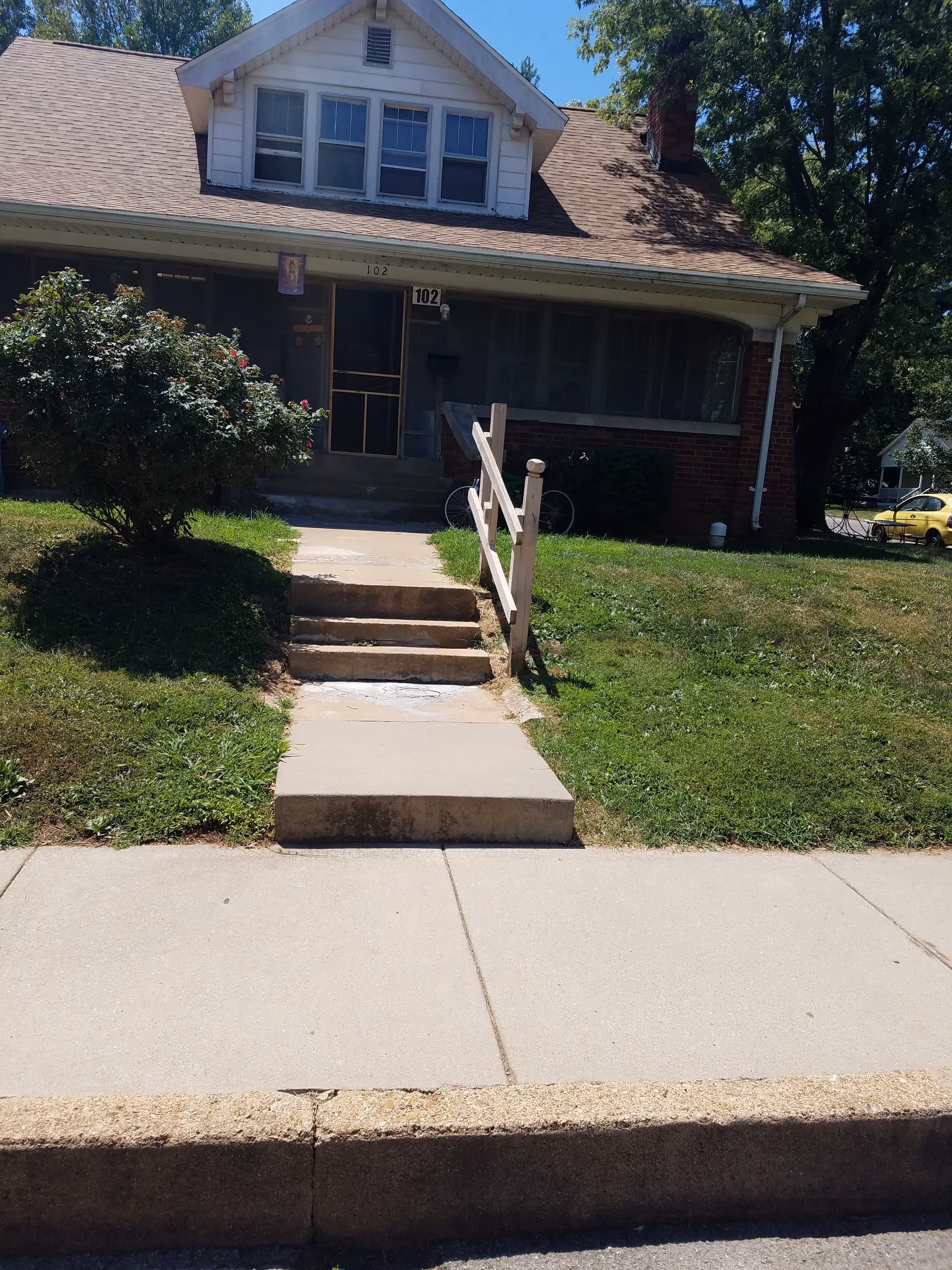 Front exterior view of a single-family house with a small porch, steps leading up to the entrance, a bush on the left side, and a bicycle partially visible behind the porch. The house has a gabled roof and several windows on the upper floor. The lawn is green and there is a sidewalk in front of the house.