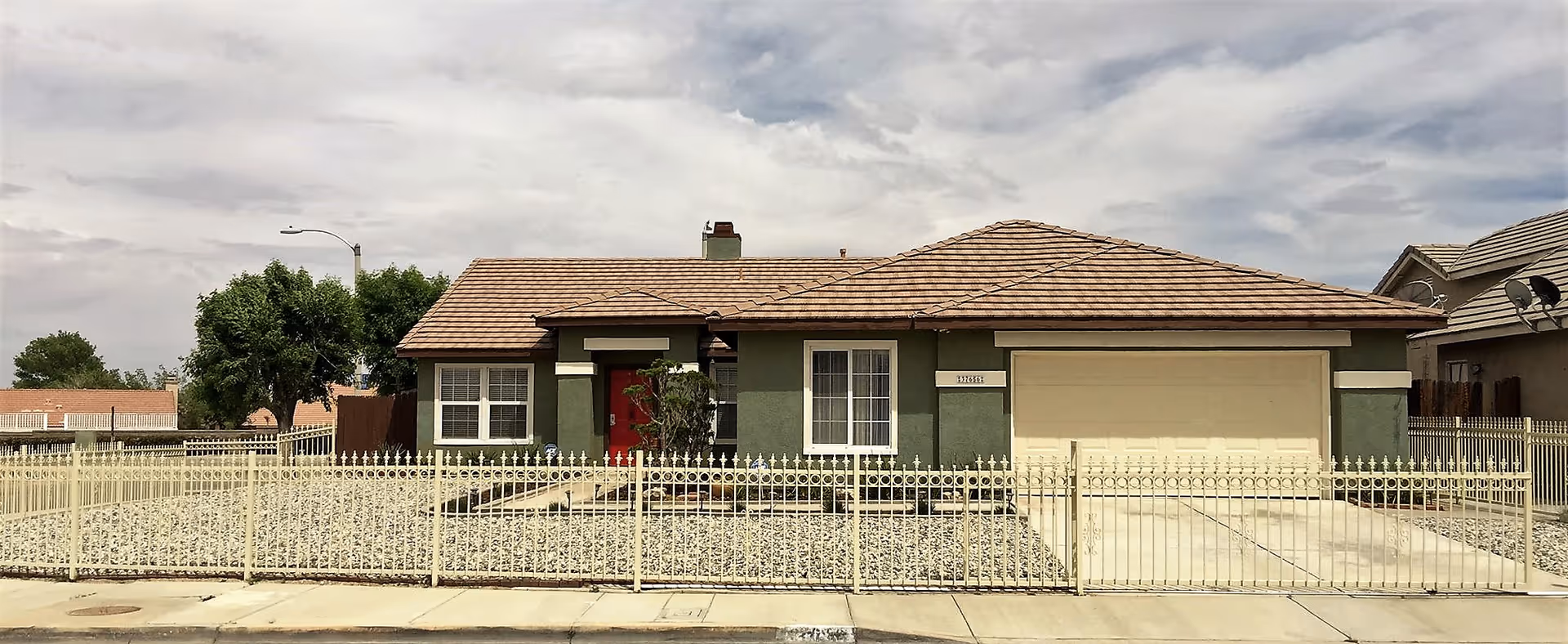 Single-story residential house with a brown tiled roof, green exterior walls, a red front door, and a two-car garage. The front yard is covered with gravel and enclosed by a decorative metal fence. There are a few trees and a cloudy sky in the background.