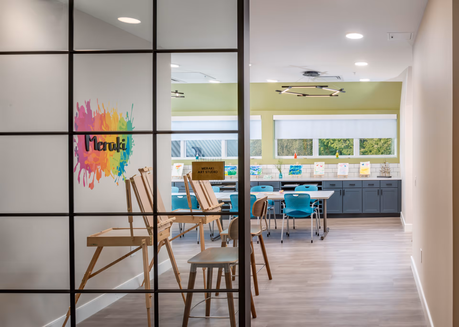 View through a glass partition into an art studio room with wooden easels, chairs, tables, and colorful paintings displayed on a counter beneath windows. The wall has a colorful splash design with the word 'Meraki'.