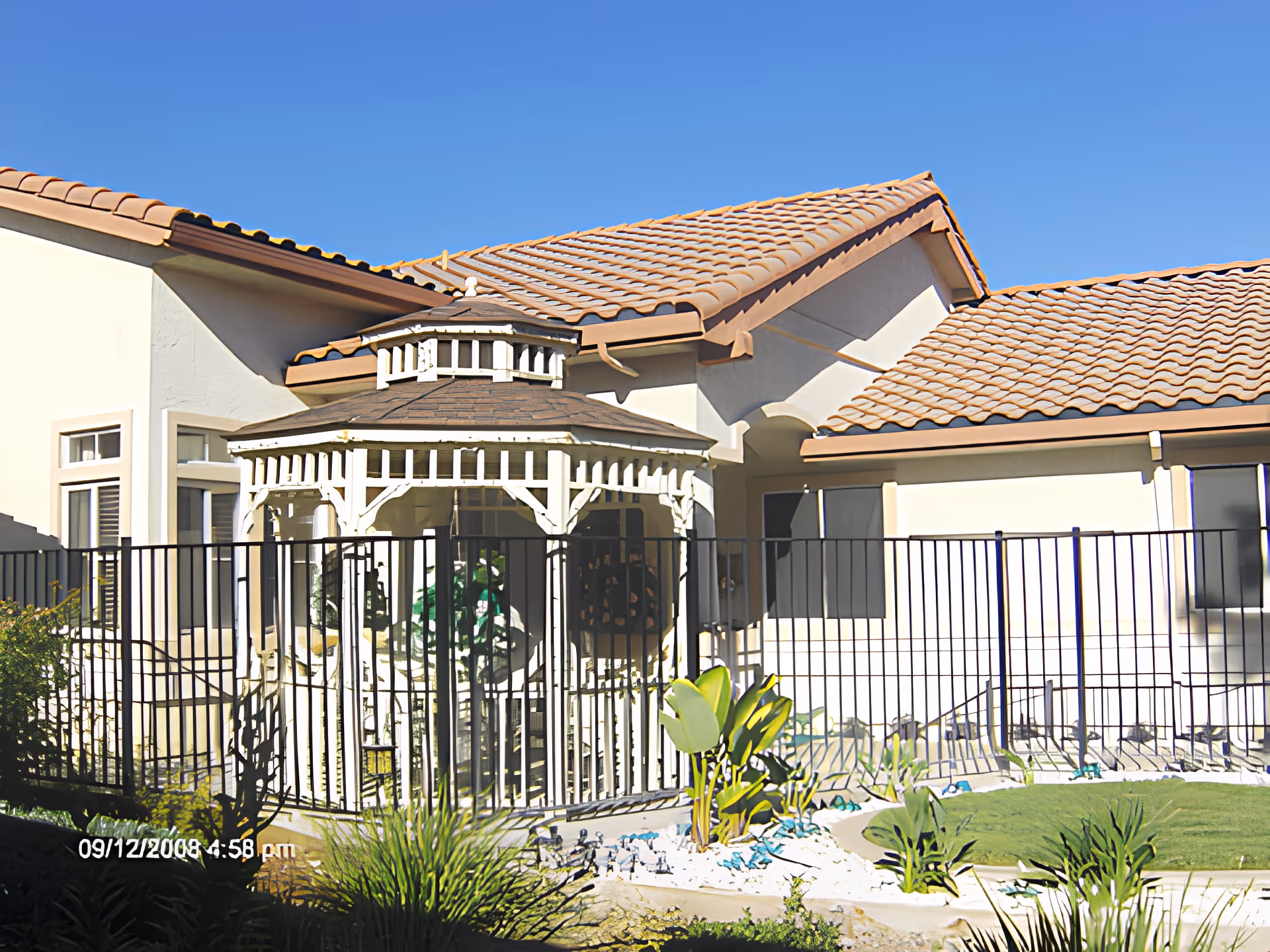 Outdoor view of a senior living facility showing a white gazebo with a brown roof enclosed by a black metal fence. The building has beige walls and a tiled roof. There are plants and a small garden area with green grass and decorative stones in front of the fence under a clear blue sky.