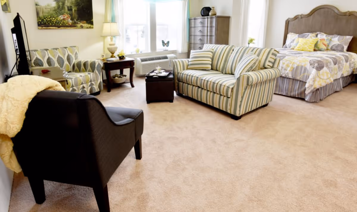 A bright and cozy room featuring a bed with patterned bedding, a striped loveseat with matching pillows, a patterned armchair, a black armchair with a yellow throw blanket, a wooden dresser, a side table with a lamp, and a window with white curtains letting in natural light.
