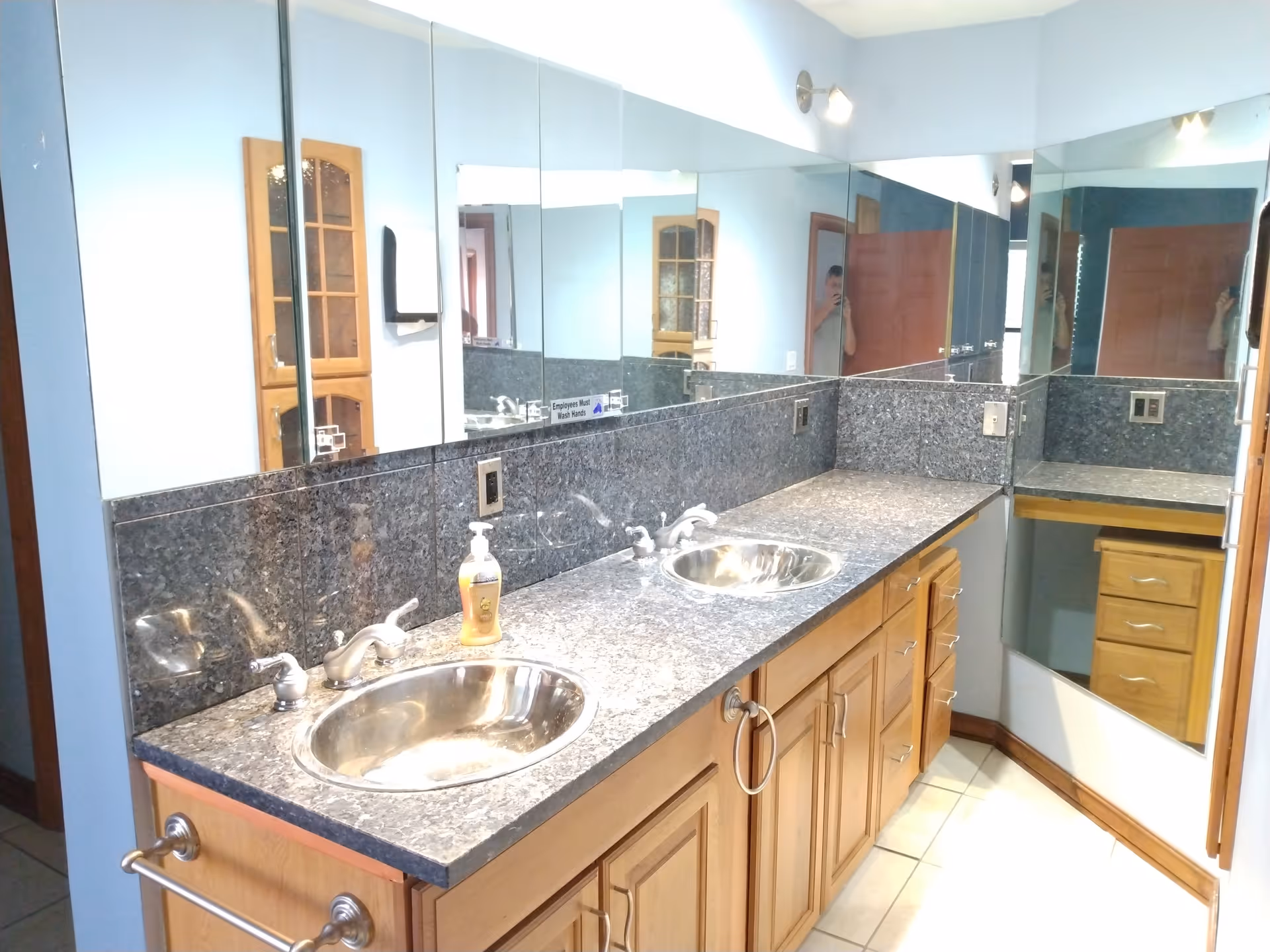A bathroom vanity area with two stainless steel sinks set in a gray granite countertop. The vanity has wooden cabinets and drawers underneath. Large mirrors cover the walls above the countertop, reflecting the room and a person taking the photo. The walls are painted light blue, and there is a towel ring and a soap dispenser on the counter.