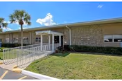 Exterior view of a single-story building with a stone facade and a covered entrance with a wheelchair ramp. There is a palm tree and well-maintained grass in front of the building under a blue sky with some clouds.