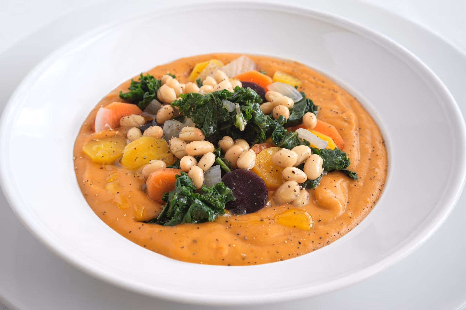 A white bowl filled with creamy orange soup topped with white beans, kale, sliced carrots, onions, and other colorful vegetables, garnished with black pepper and a drizzle of oil.