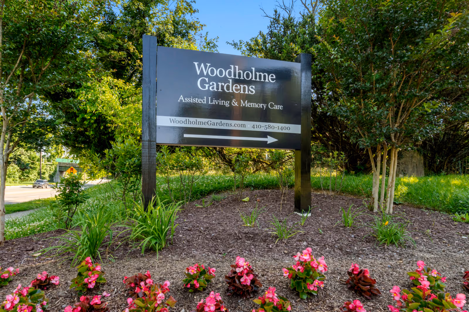 A sign for Woodholme Gardens, an assisted living and memory care facility, surrounded by greenery and flowers.