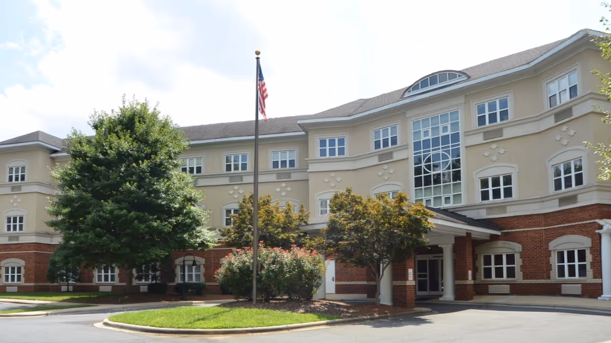 Front exterior of a multi-story senior living facility with a covered entrance, flagpole, and landscaped trees and shrubs.
