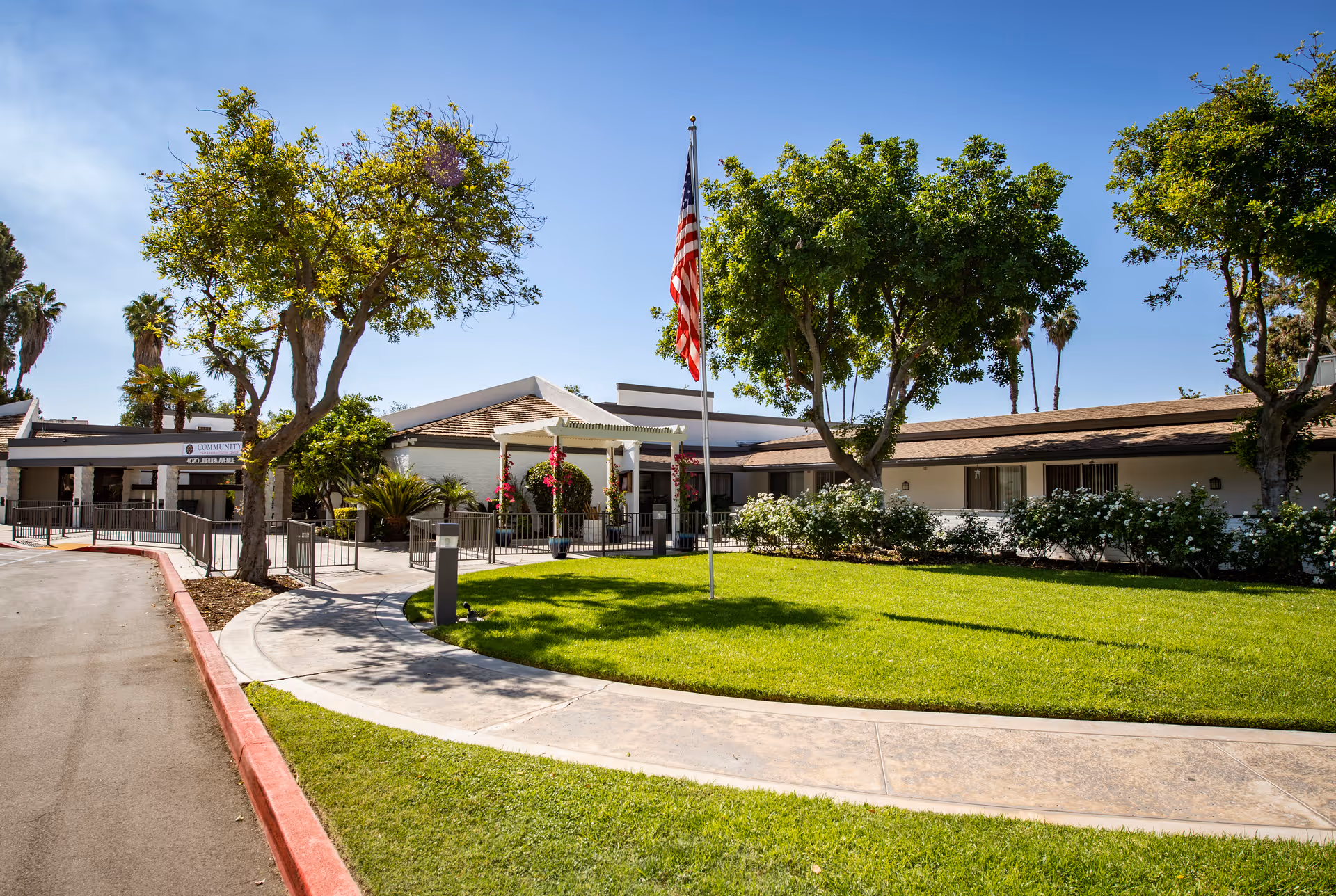 Exterior view of Community Care & Rehabilitation Center showing a single-story building with a well-maintained lawn, trees, and an American flag on a flagpole in front. The sky is clear and blue.