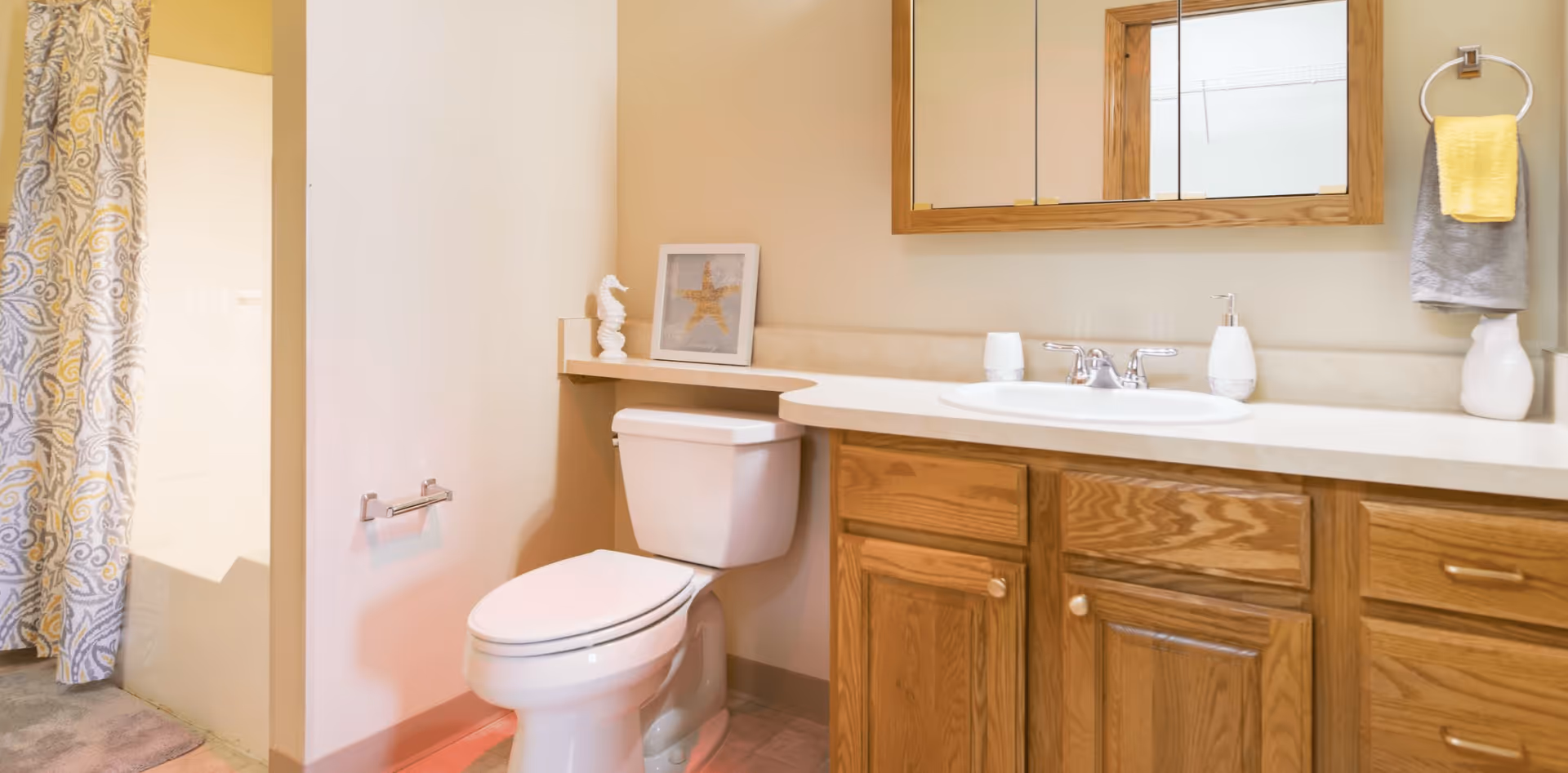 A bathroom with a white toilet, wooden cabinets with a beige countertop, a sink with a silver faucet, a wall-mounted mirror with a wooden frame, a towel ring holding a gray and yellow towel, a decorative white seahorse, and a framed starfish picture. A shower with a patterned curtain is partially visible on the left.