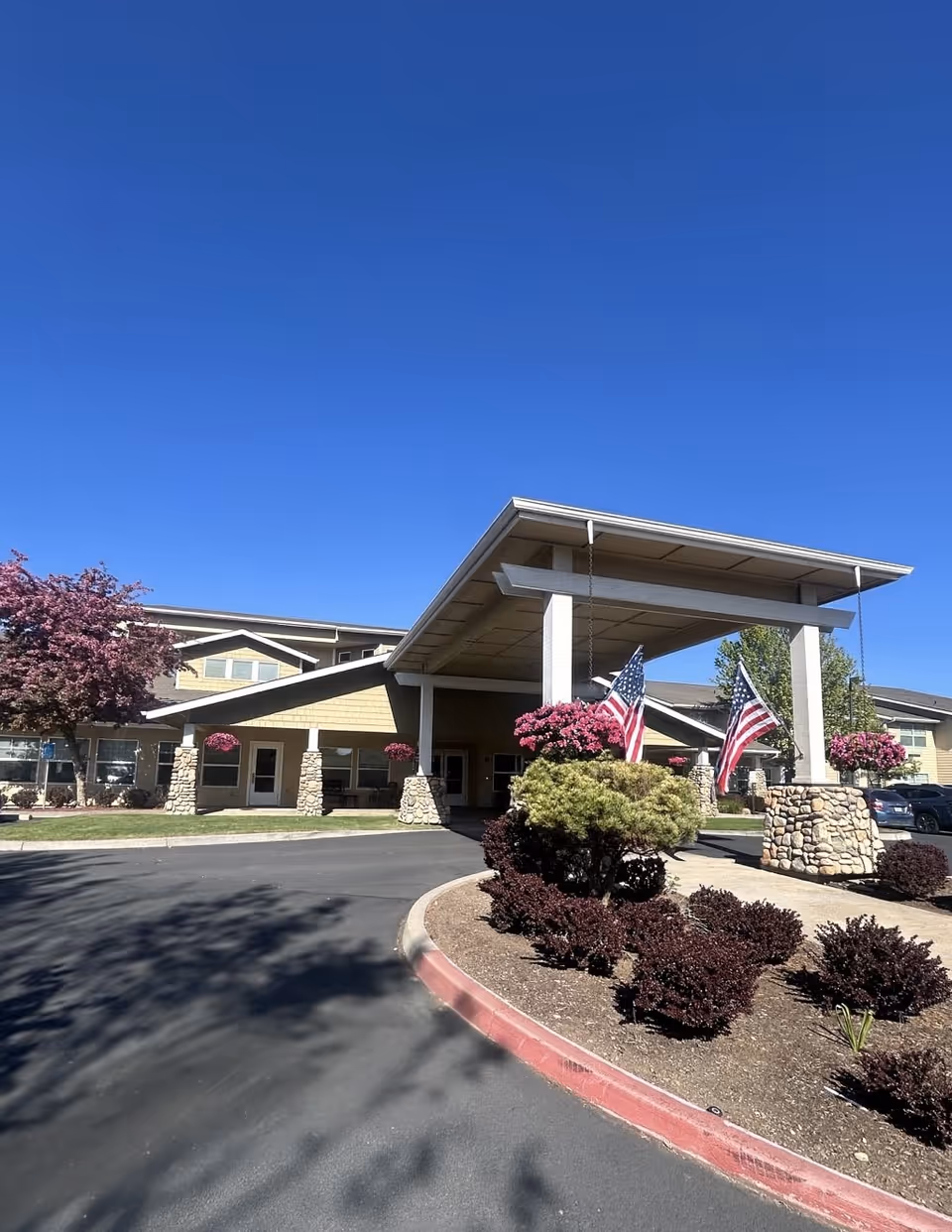 Front entrance and porte-cochère of a senior living building with American flags, hanging flower baskets, and landscaped driveway under a clear blue sky.