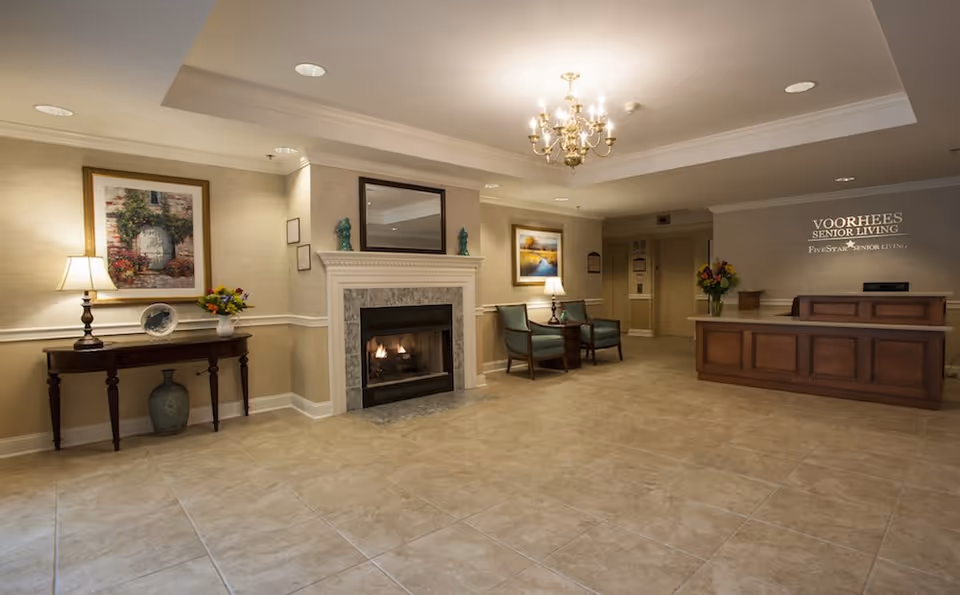 A spacious and warmly lit senior living facility lobby with a tiled floor, a fireplace with a mirror above it, two armchairs with a side table and lamp, a wooden console table with a lamp, flowers, and decorative items, and a reception desk with a flower arrangement. The wall behind the reception desk displays the text 'Voorhees Senior Living Five Star Senior Living'.