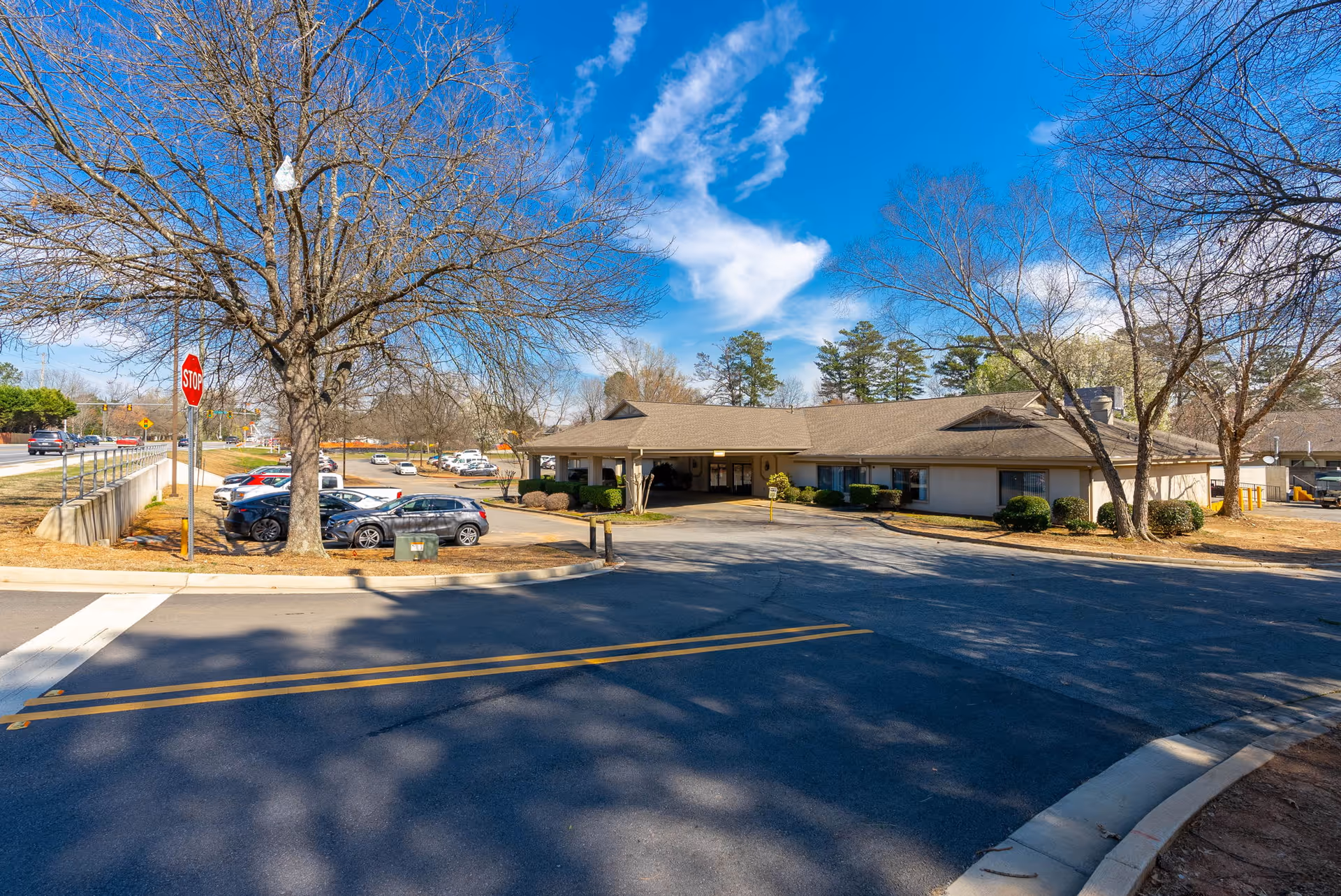 Front exterior of a single-story healthcare building with a drive-through entrance, parking lot, and leafless trees under a blue sky.