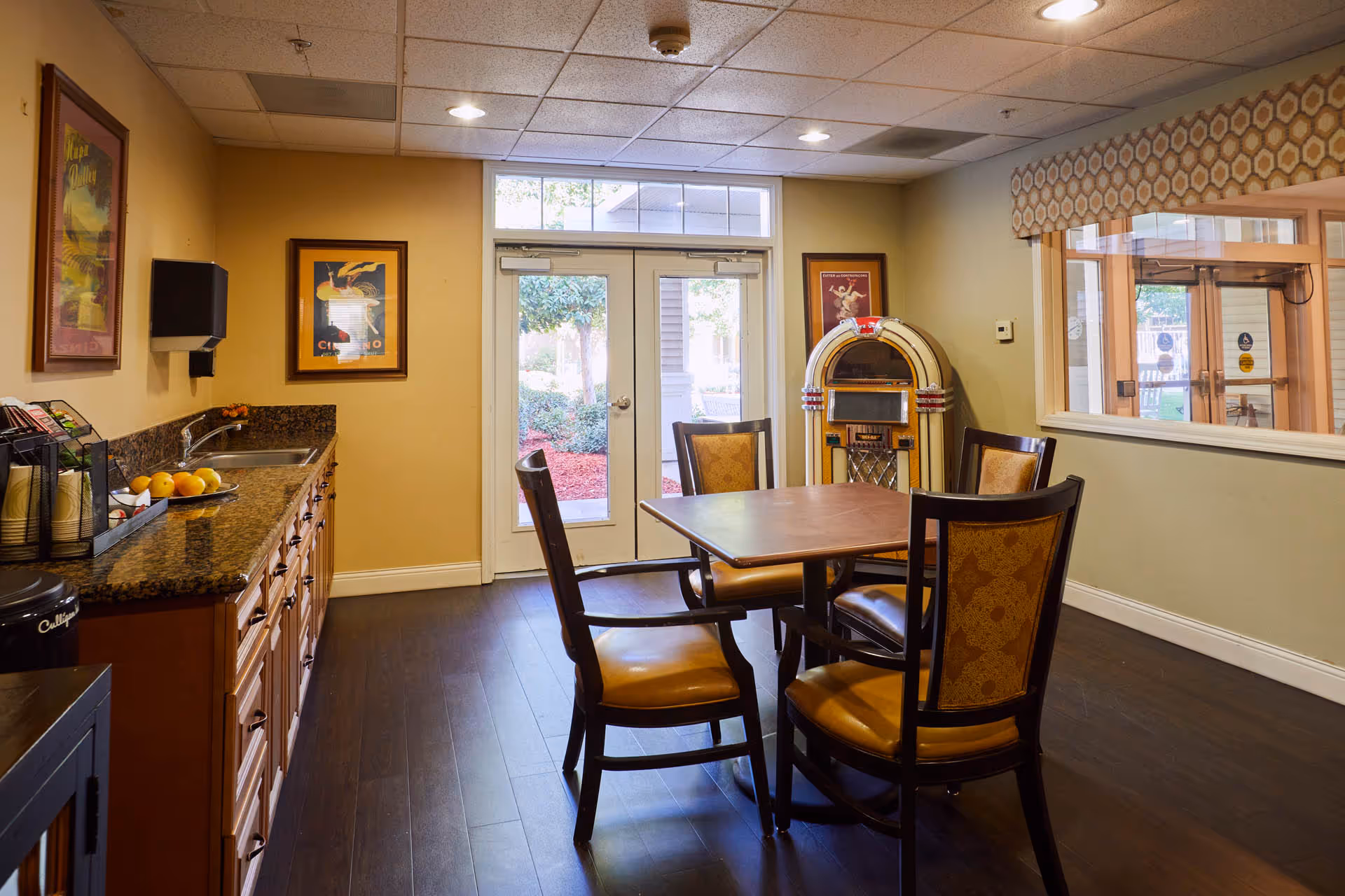 Small common room with a table and four chairs, a kitchenette along the left wall, and a jukebox near glass doors.