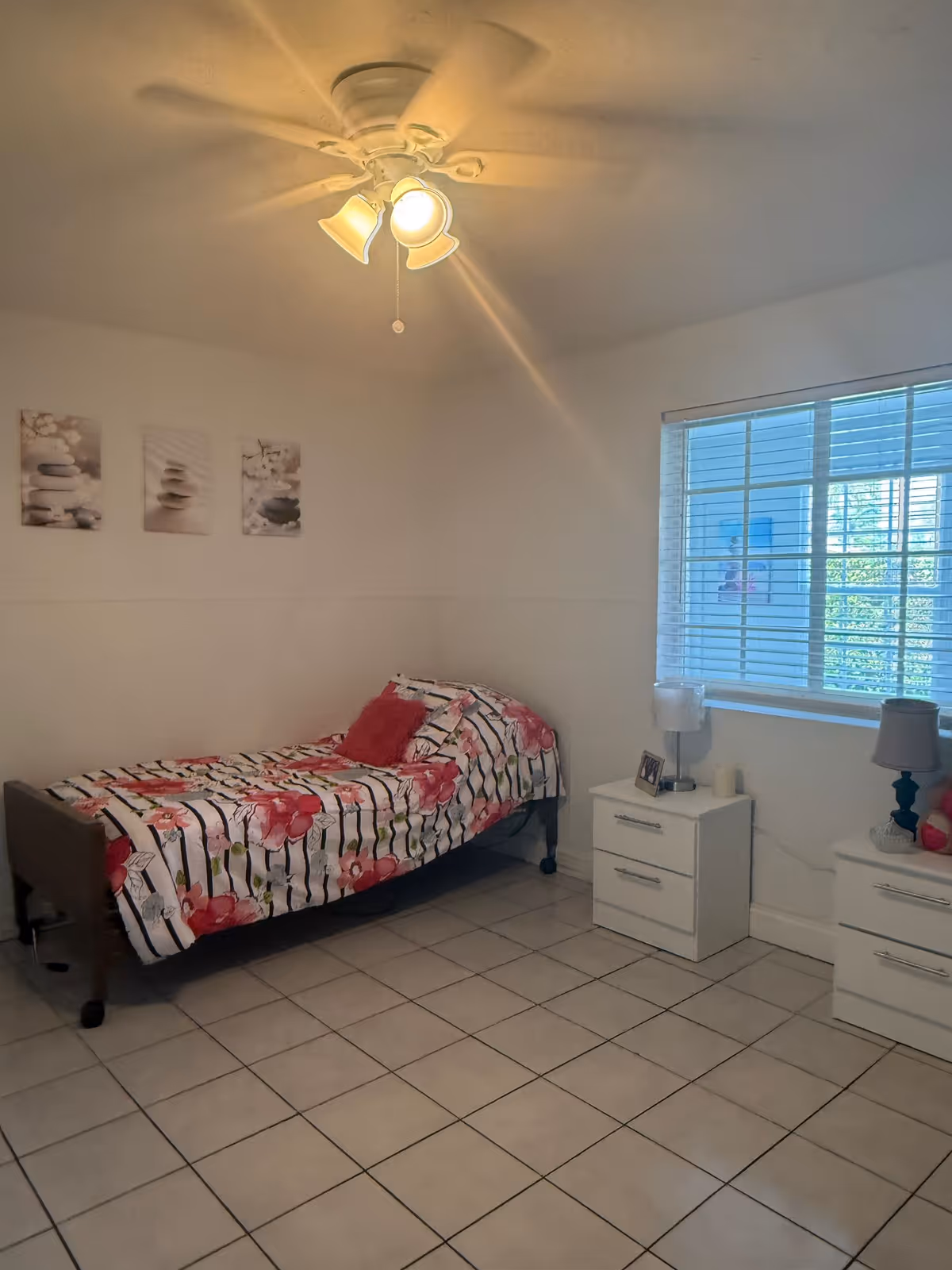 A small bedroom with a single bed covered in a floral patterned bedspread with pink and red flowers. There are two white nightstands next to the bed, each with a lamp and some small decorative items. The room has white tiled flooring, white walls, a ceiling fan with lights, and a window with white blinds letting in natural light. Three small framed pictures hang on the wall above the bed.