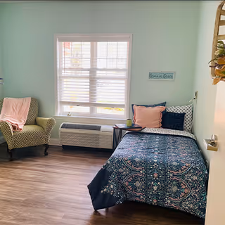Small bright bedroom with a single bed with patterned bedding, an armchair, and a window with blinds.