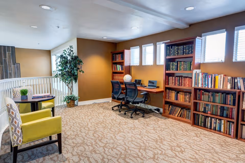 A cozy reading and computer area in a senior living facility with two bookshelves filled with books, two black office chairs at a wall-mounted desk with two laptops, a globe, and a potted plant in the corner. There are also two patterned armchairs and a small round table with a plant and magazines. The room has beige walls, carpeted floor, and several windows letting in natural light.