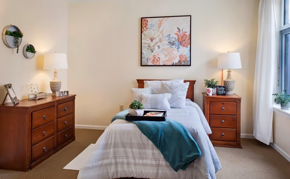 A neatly made bed with white bedding and multiple pillows in a bedroom. There is a teal throw blanket and a tray with a small plant on the bed. On either side of the bed are wooden nightstands with matching lamps and decorative items. A floral painting hangs above the bed, and there is a window with white curtains on the right side letting in natural light. A wooden dresser with framed photos and decorative plants is on the left side of the room.