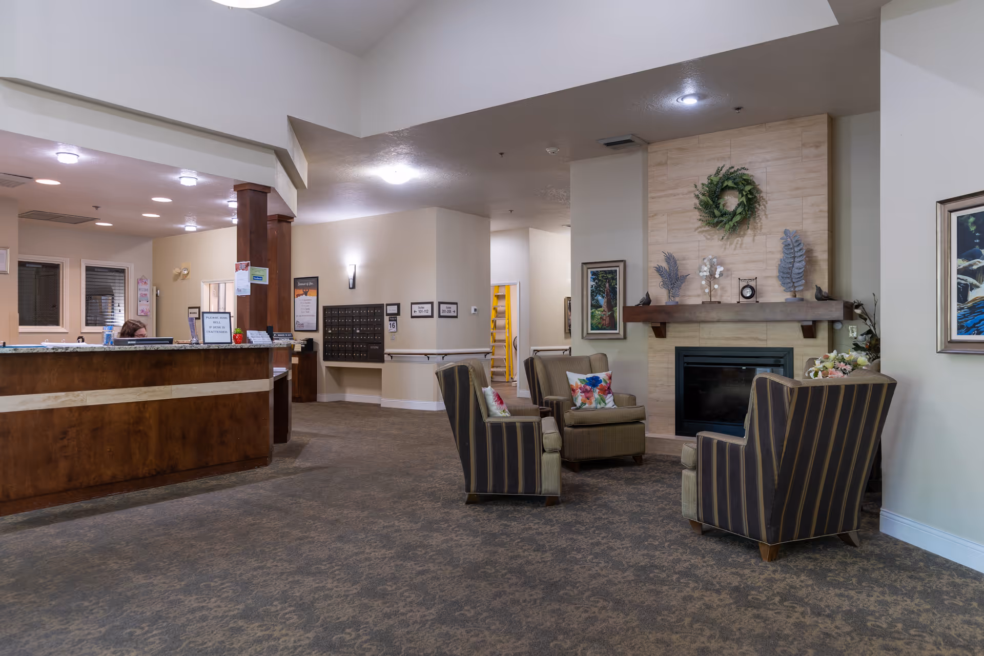 Interior view of a senior living facility lobby with a wooden reception desk on the left, a seating area with four striped armchairs and colorful pillows arranged around a fireplace decorated with a wreath and ornaments, and a carpeted floor.