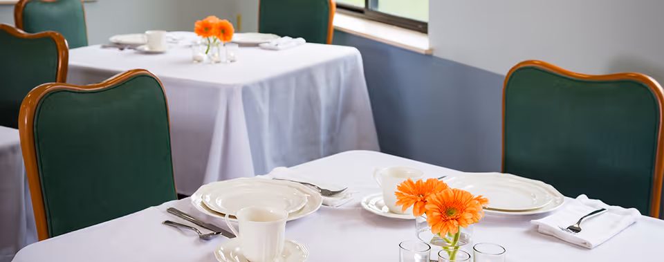 A dining area with tables covered in white tablecloths, set with white plates, cups, silverware, and napkins. Each table has a small vase with orange flowers. Green cushioned chairs with wooden frames surround the tables. A window is visible in the background.