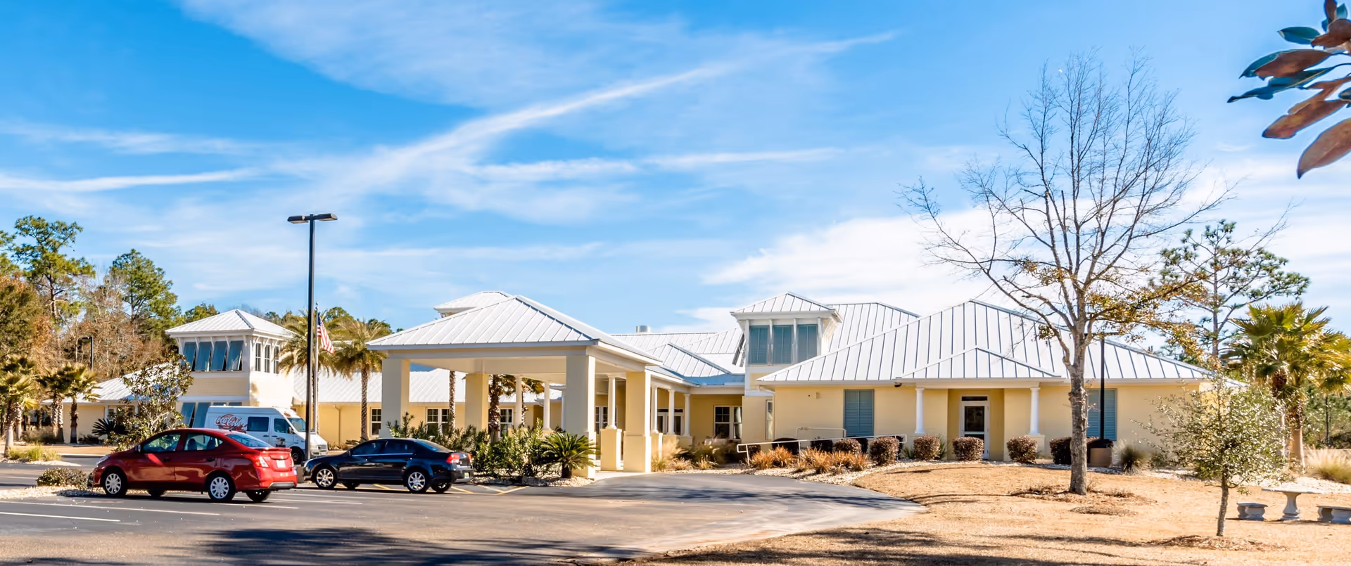 Front exterior of a single-story memory care facility with a covered entrance, parked cars and landscaping under a blue sky.