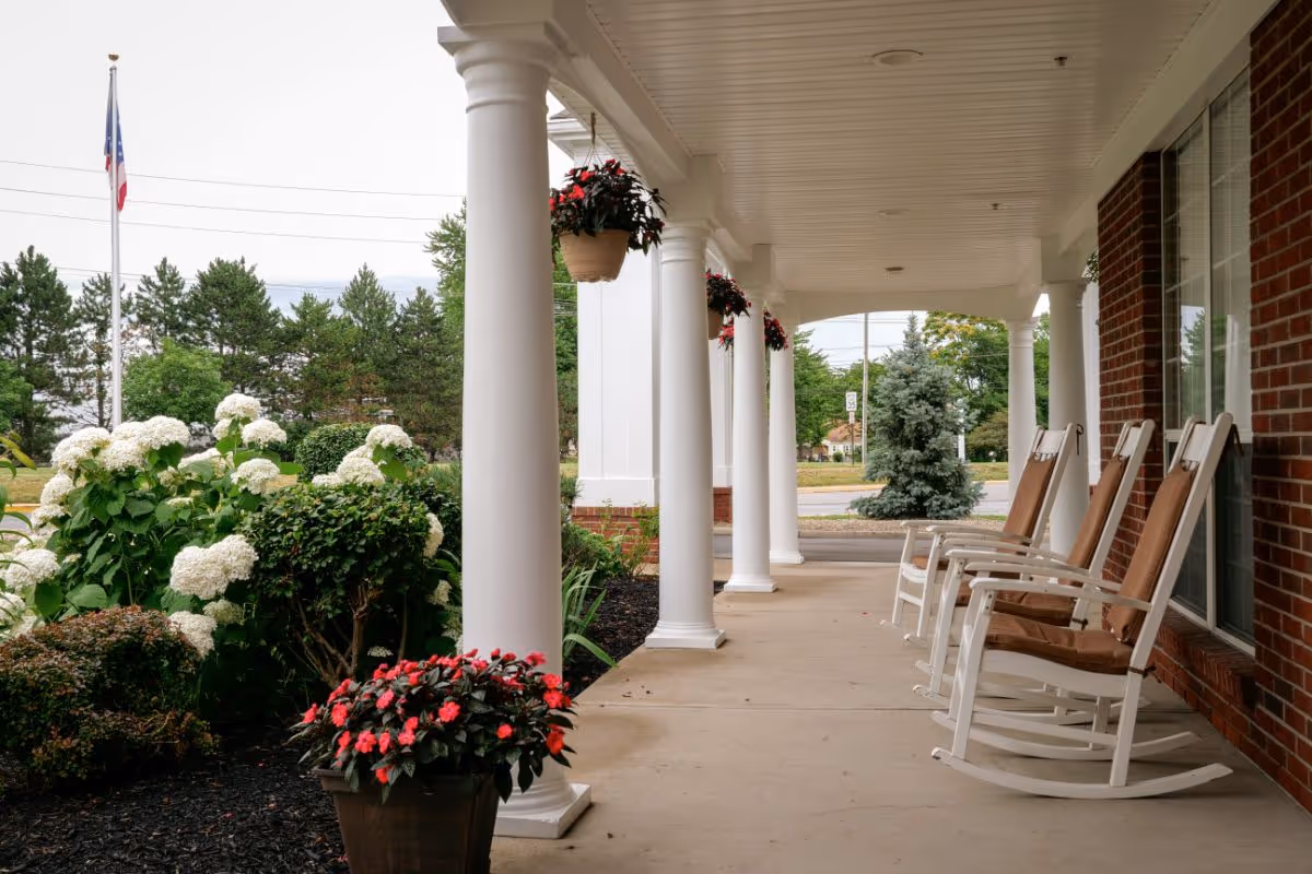 A covered porch with white columns and four white rocking chairs with brown cushions lined up against a brick wall. There are hanging flower pots with red flowers and potted plants with white and red flowers near the columns. In the background, there are trees, a flagpole with an American flag, and a street.
